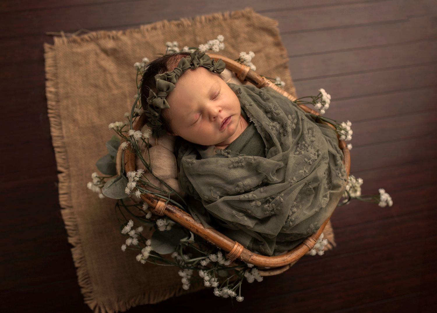Sleeping newborn wrapped in gray fabric surrounded by delicate flowers in a rustic basket.