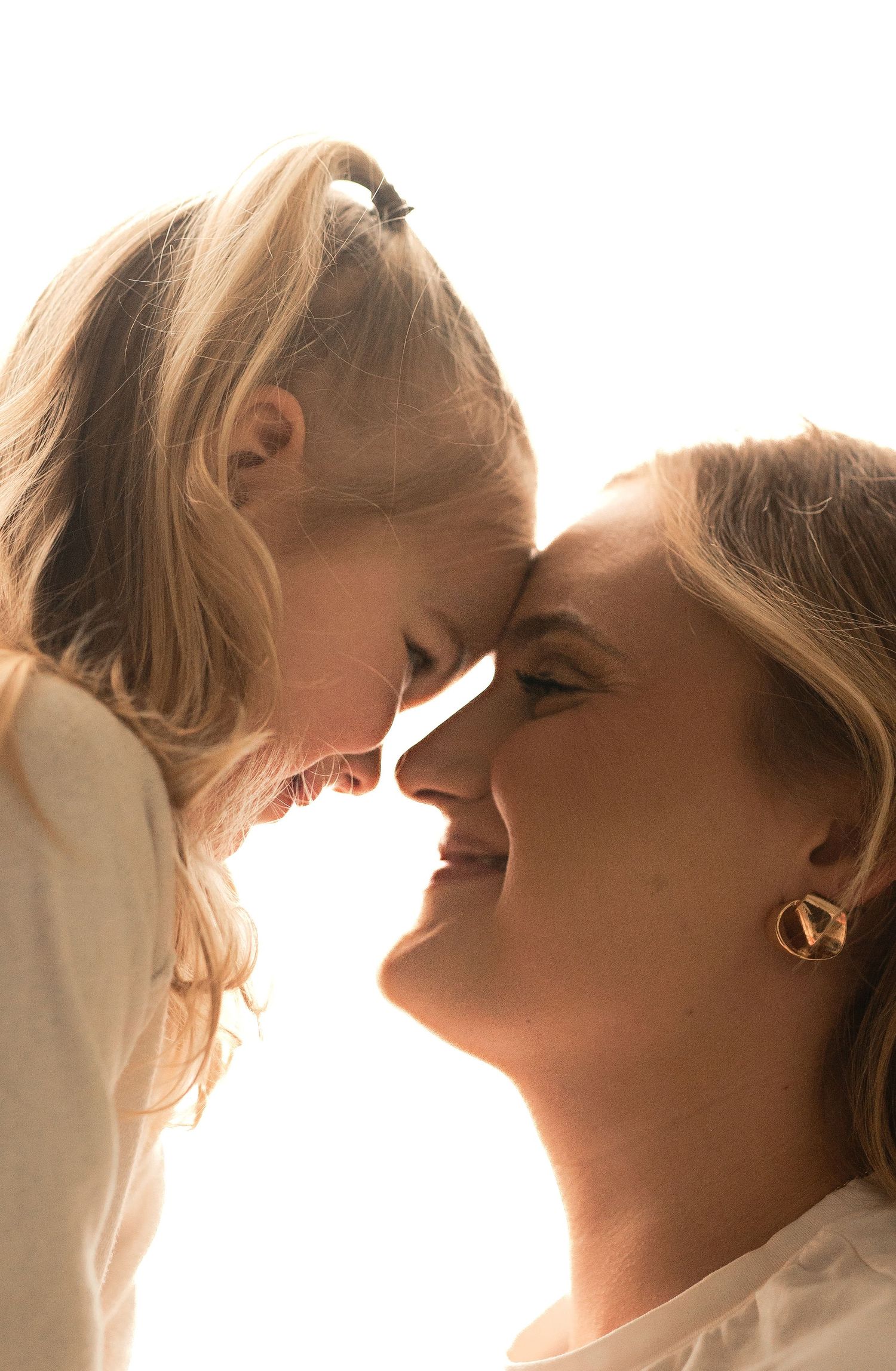 Mother and daughter touching foreheads smiling in bright light.
