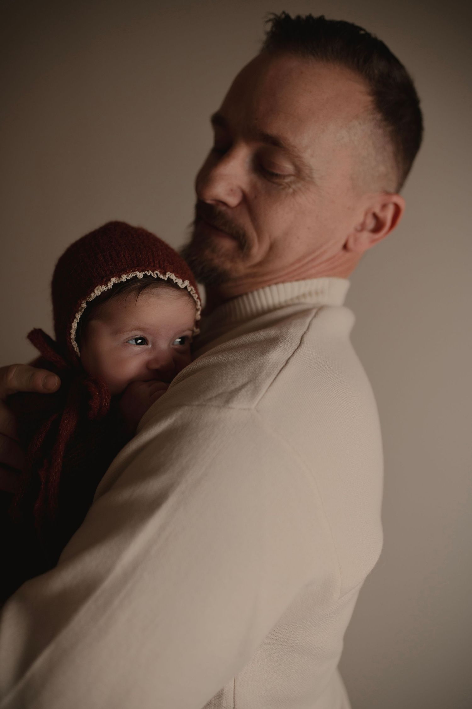 Parent in cream sweater holding small baby wearing burgundy knit hat in soft lighting.