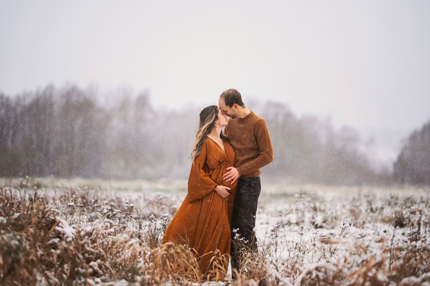 A romantic couple embraces in a snowy winter field wearing warm clothing at sunset.