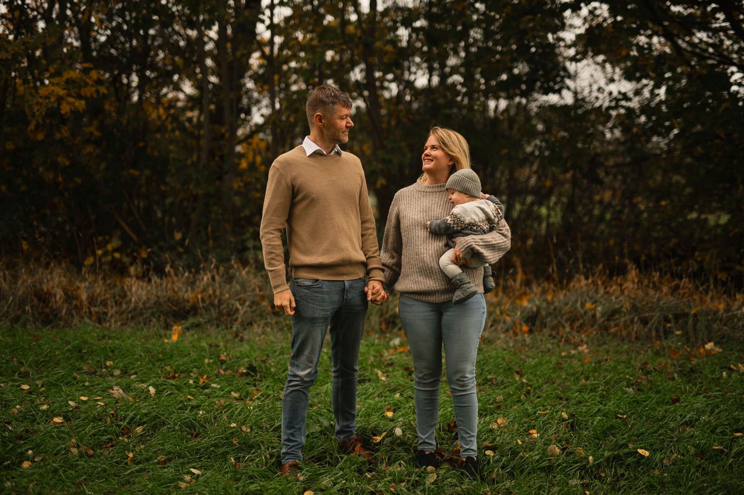A couple in cozy sweaters stands together on grassy autumn ground with trees in the background.