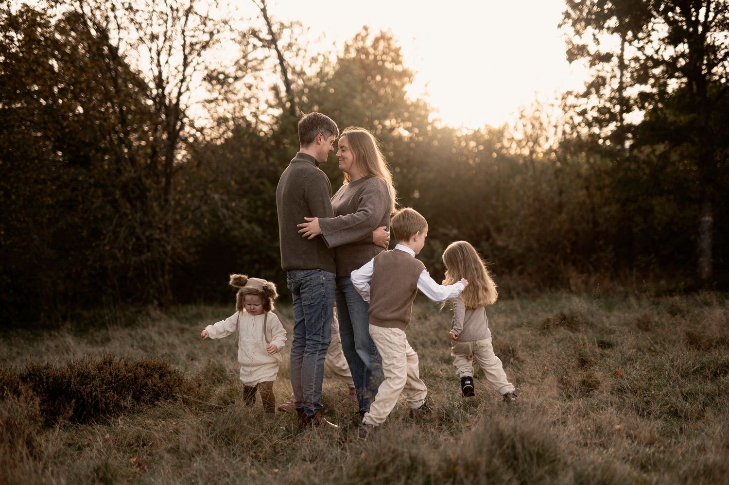 Family of five embracing at golden hour in autumn field with two young children playing around parents.