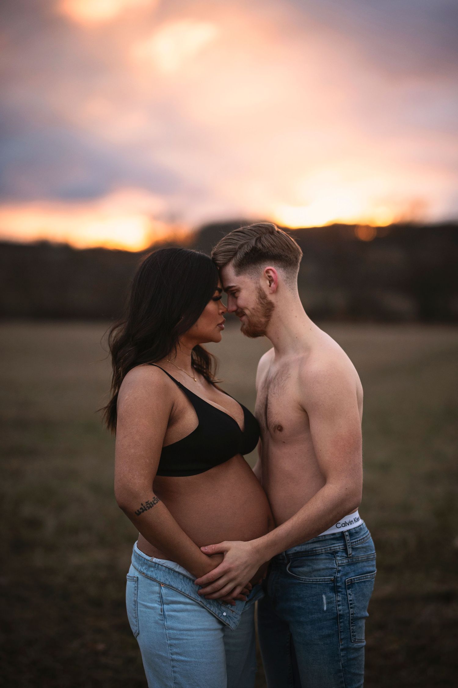 A couple shares a tender moment during their sunset maternity photoshoot in an open field.