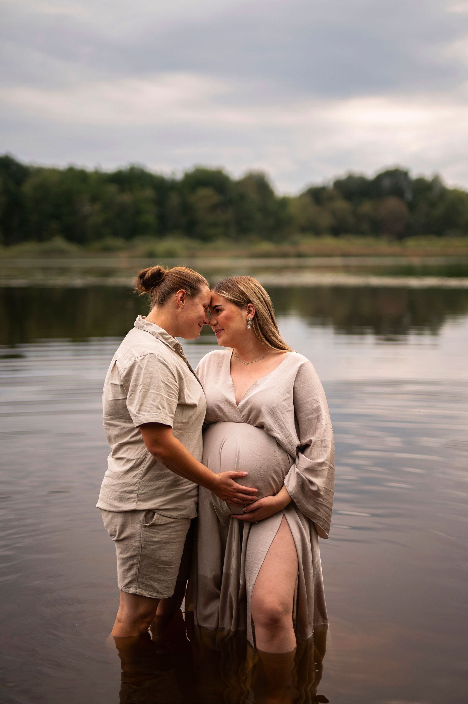 Couple standing in a lake, woman in flowing maternity dress cradling her bump, partner embracing her in a romantic outdoor shoot.
