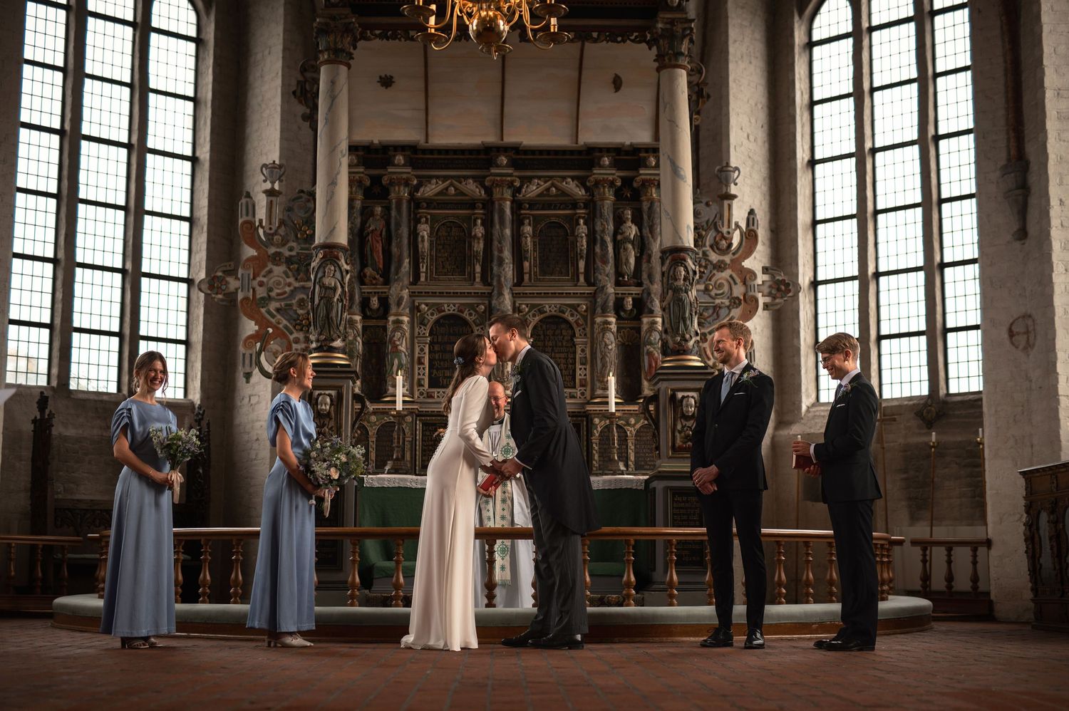 Couple exchanging vows at ornate church altar with bridesmaid and groomsman witnesses during intimate wedding ceremony.