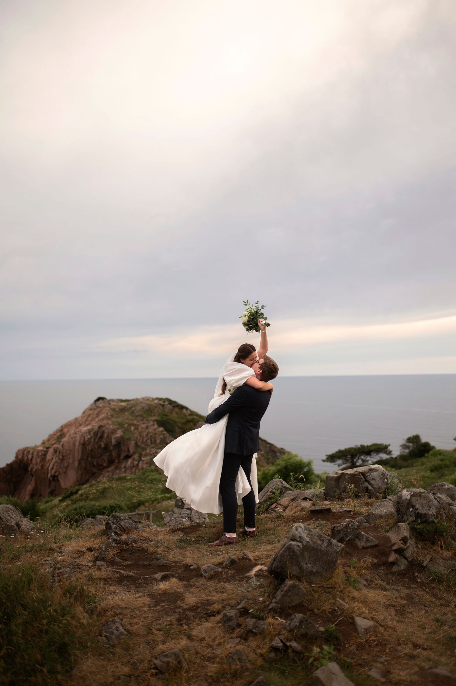 Groom lifting bride with bouquet on rocky coastal cliff at dusk during outdoor wedding photo shoot.  Kullaberg elopment