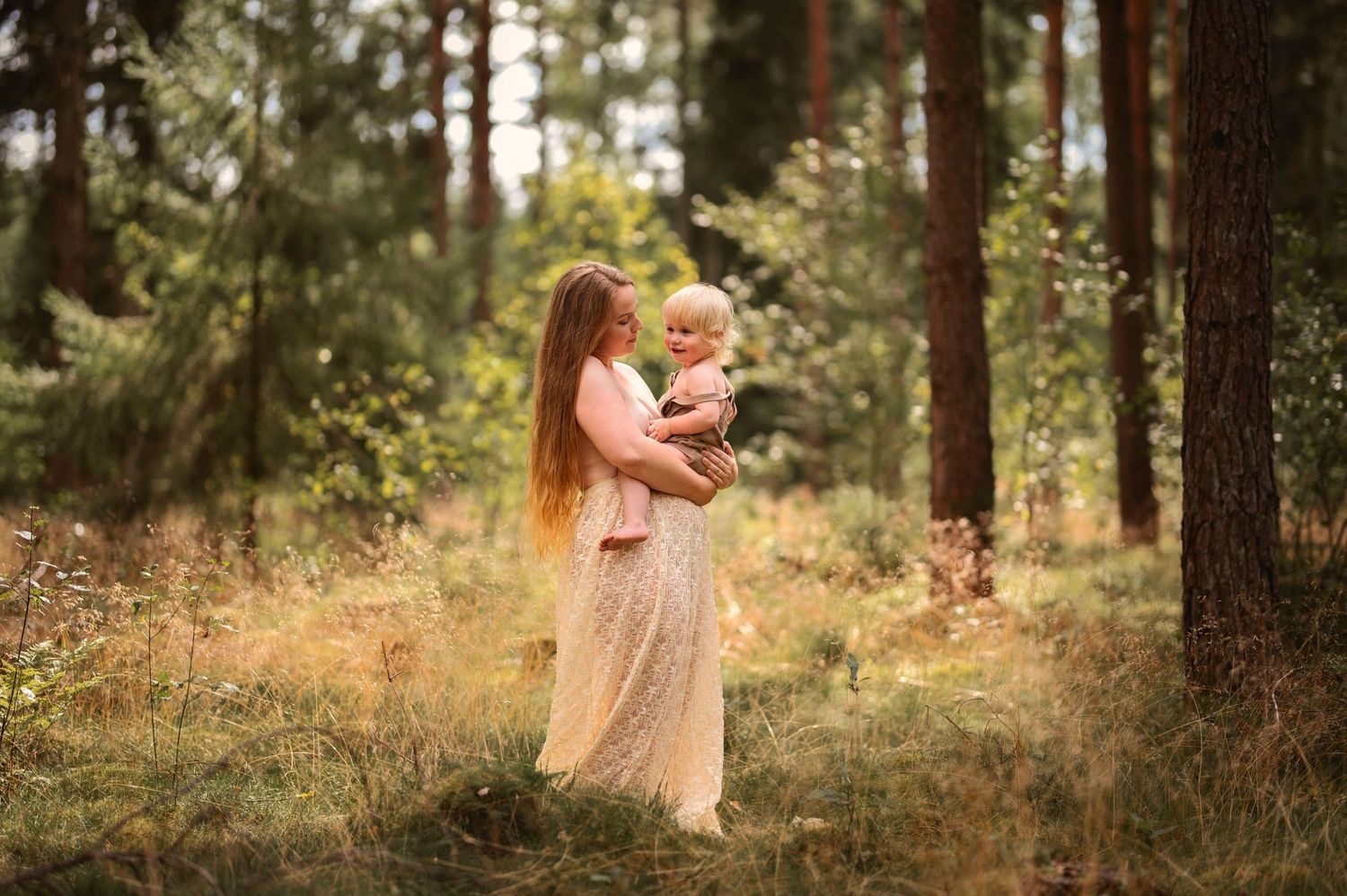 Moderskapsfotografering i Skåne med förälder som håller ett litet barn i spetsklänning i drömlik skogsmiljö.