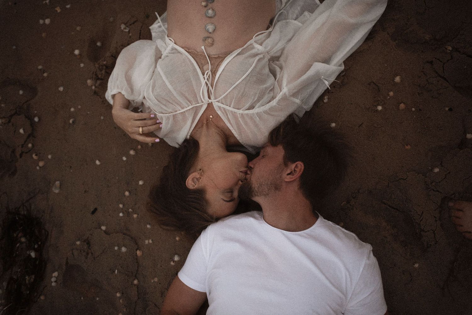 Couple sharing a romantic kiss while lying on a sandy beach from above.