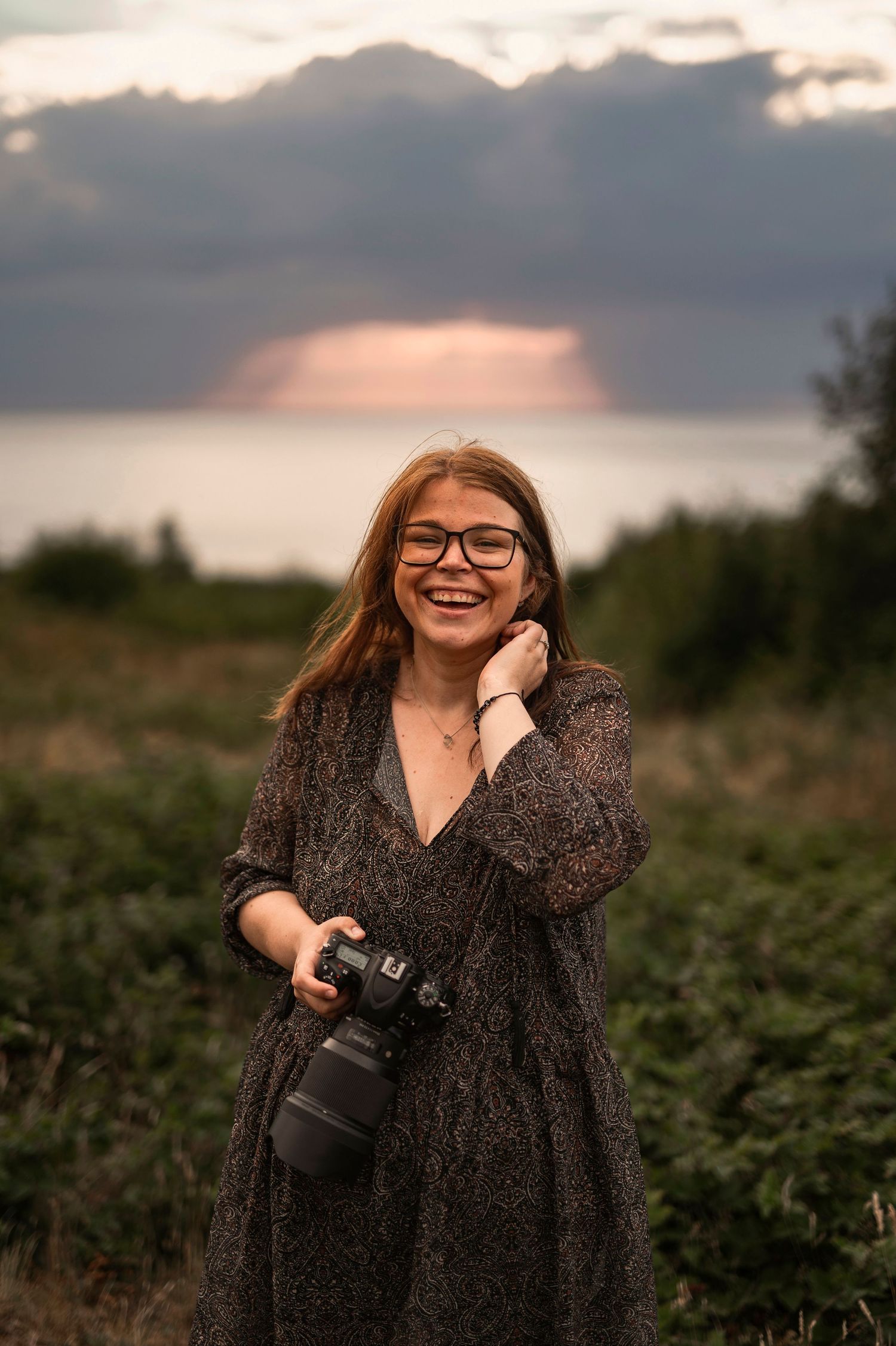 at Hovs Hallar. Smiling redhead woman with glasses holding a DSLR camera outdoors at sunset in a floral dress.