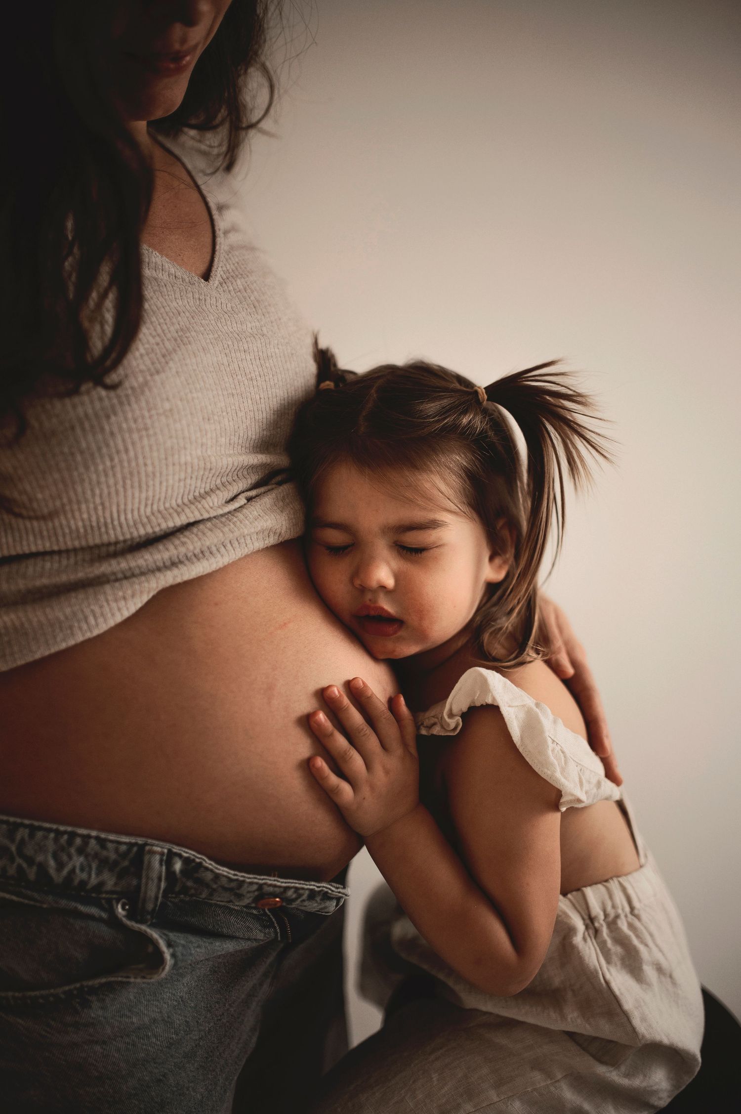 A young toddler girl rests her cheek on her pregnant mother's bare belly, gently placing her hand on it.