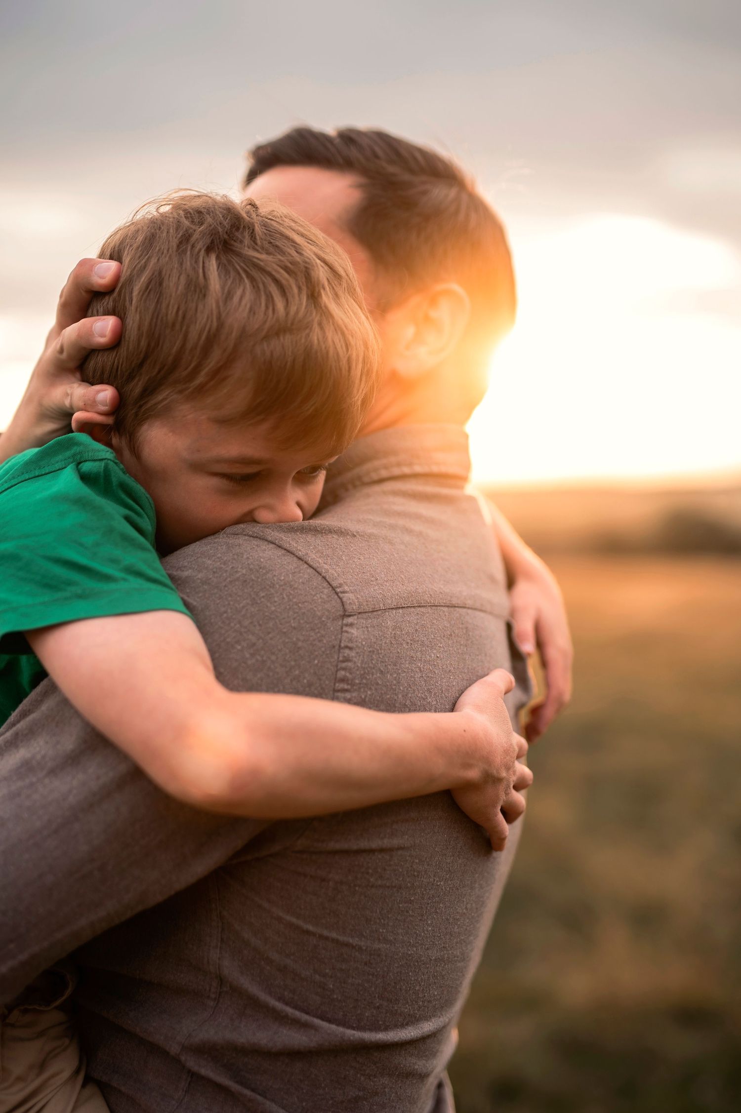 Familjefotografering i Skåne med pojke som kramar sin pappa i gyllene kvällsljus på fält.