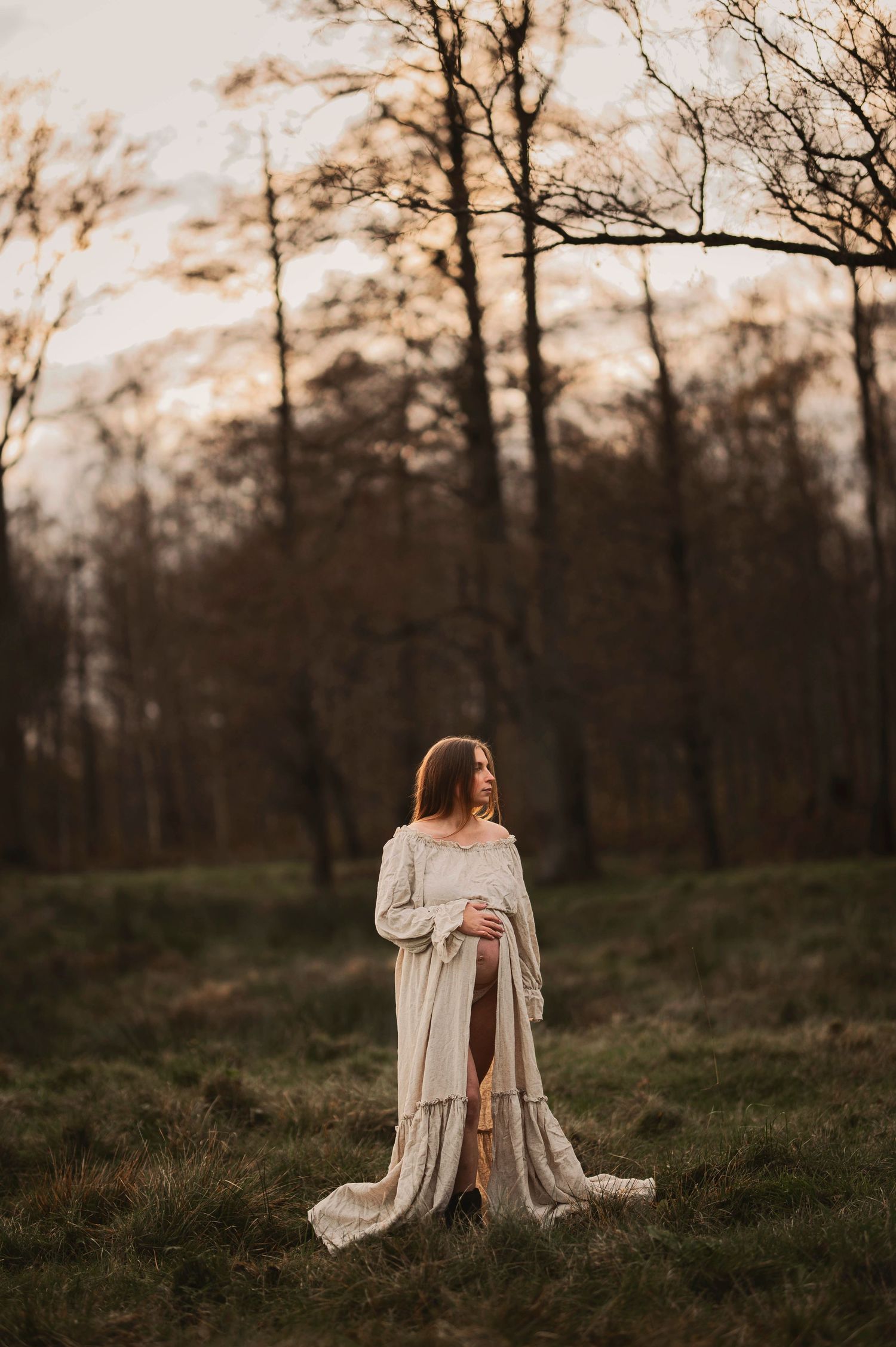 Gravidfotografering i Skåne med gravid kvinna i linneklänning i skogsglänta i gyllene kvällsljus.