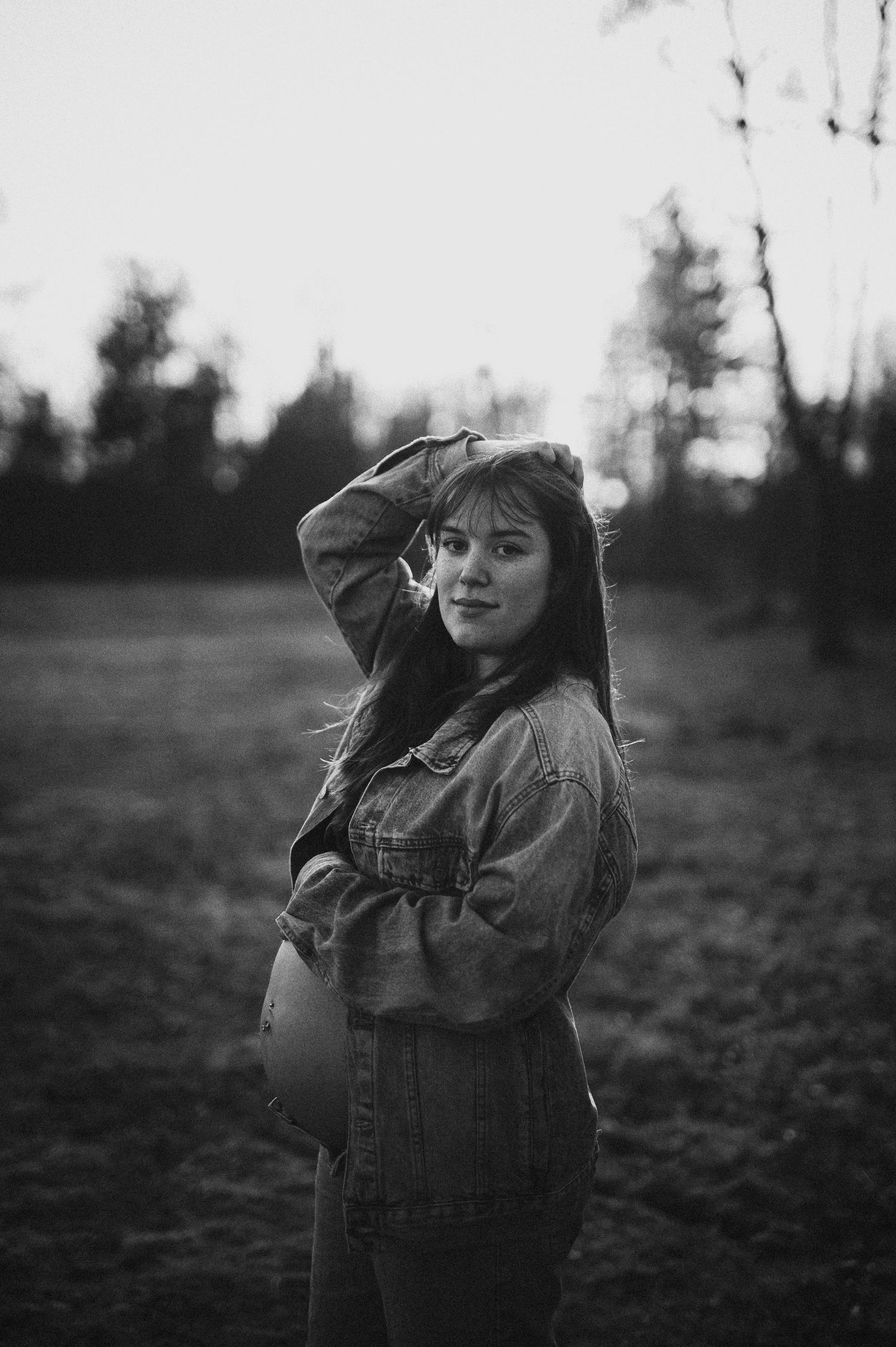 Svartvit gravidfotografering i Skåne med gravid kvinna i jeansjacka i fält i kvällsljus.