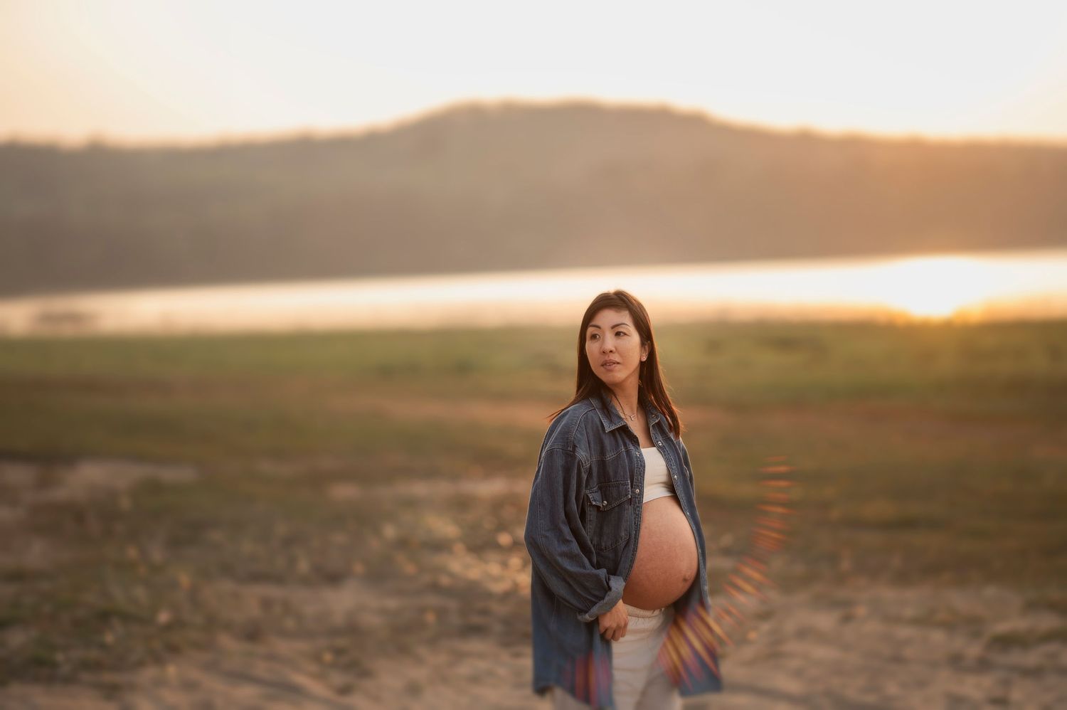 Gravid kvinna i jeansjacka fotograferad i ett öppet landskap under en gravidfotografering i Skåne i kvällsljus