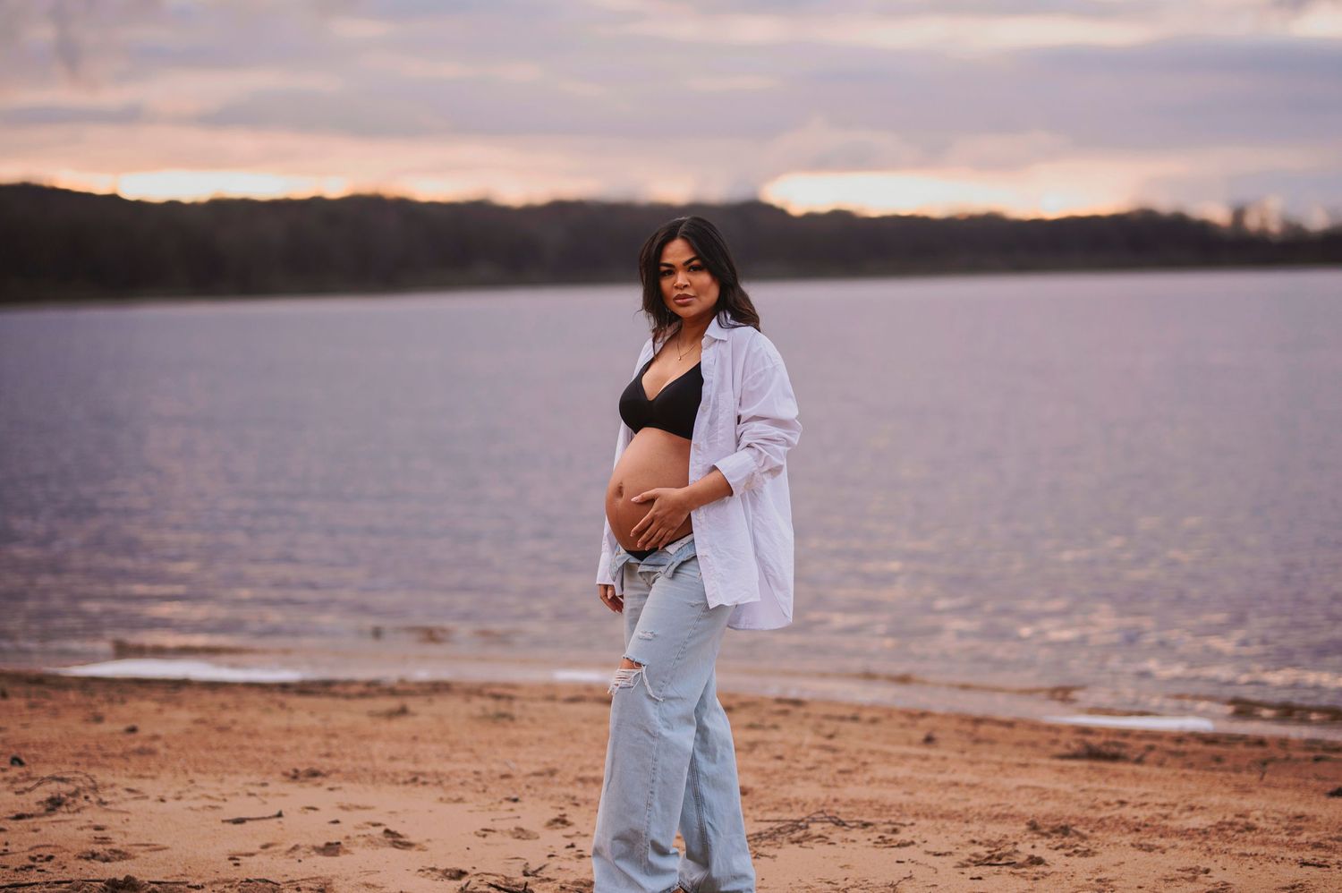Pregnant woman in casual attire poses on beach during golden hour.