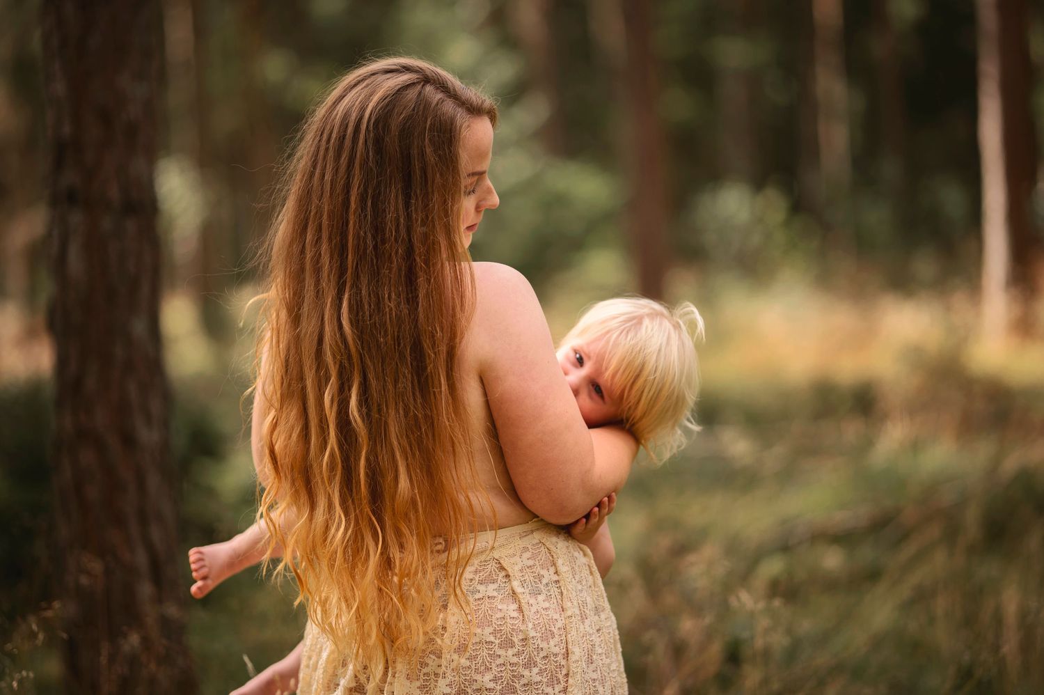 Moderskapsfotografering i Skåne med förälder som håller ett litet barn i spetsklänning i drömlik skogsmiljö.