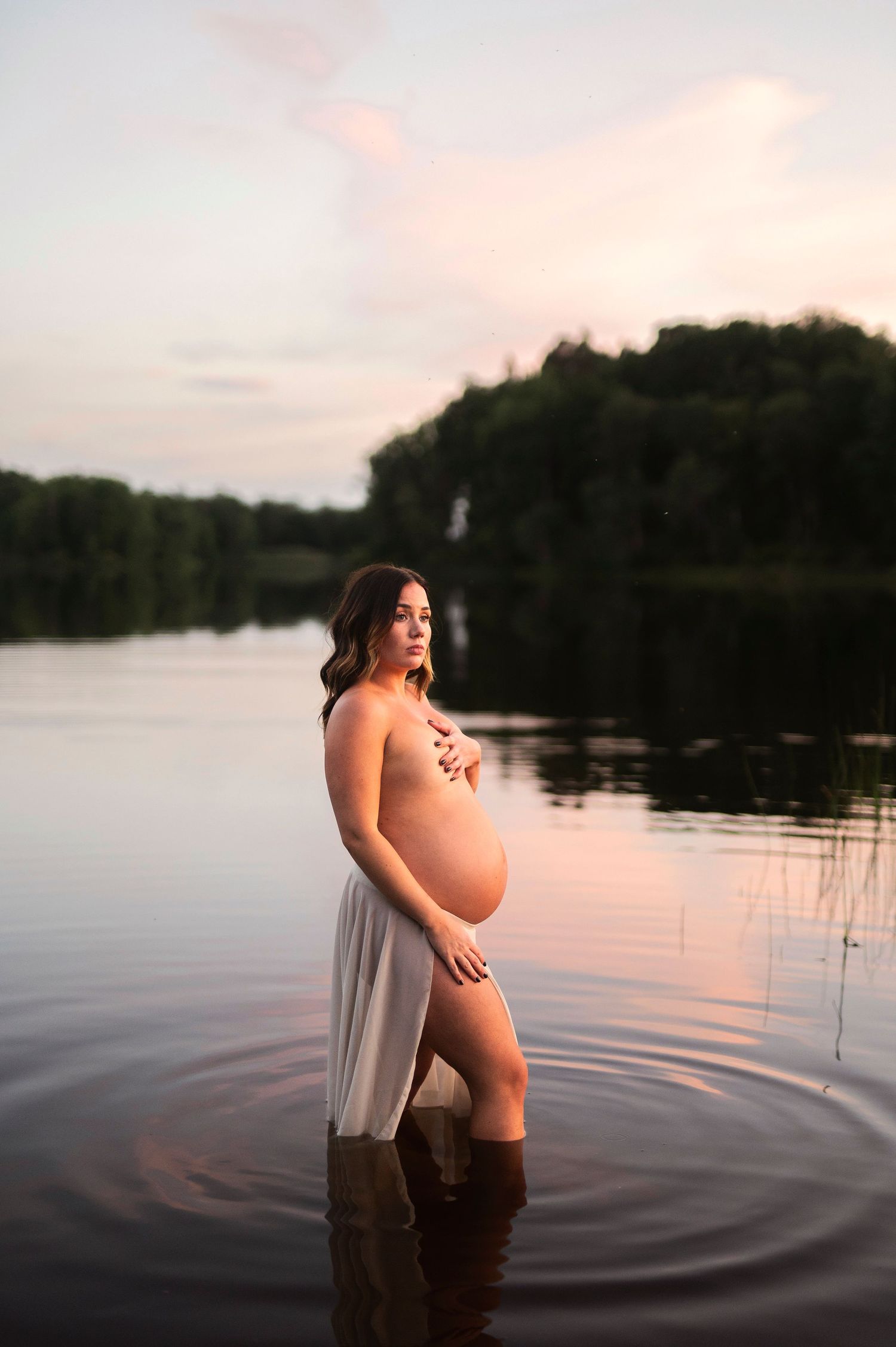 Gravidfotografering i Skåne med blivande mamma i lugnt sjövatten vid solnedgång i natur.