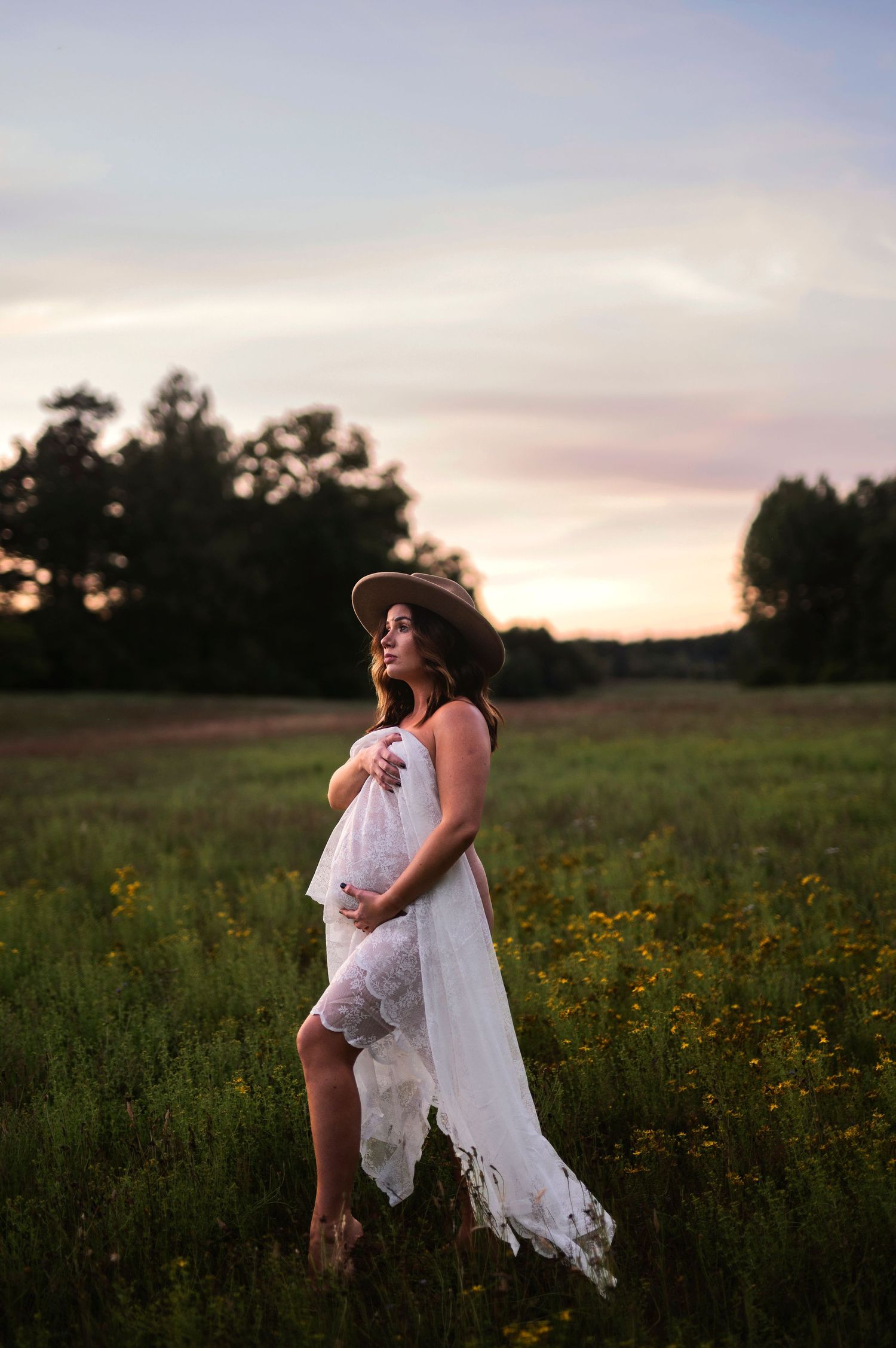 Gravidfotografering i Skåne med blivande mamma i vit spetsklänning bland vildblommor i skymningsljus.