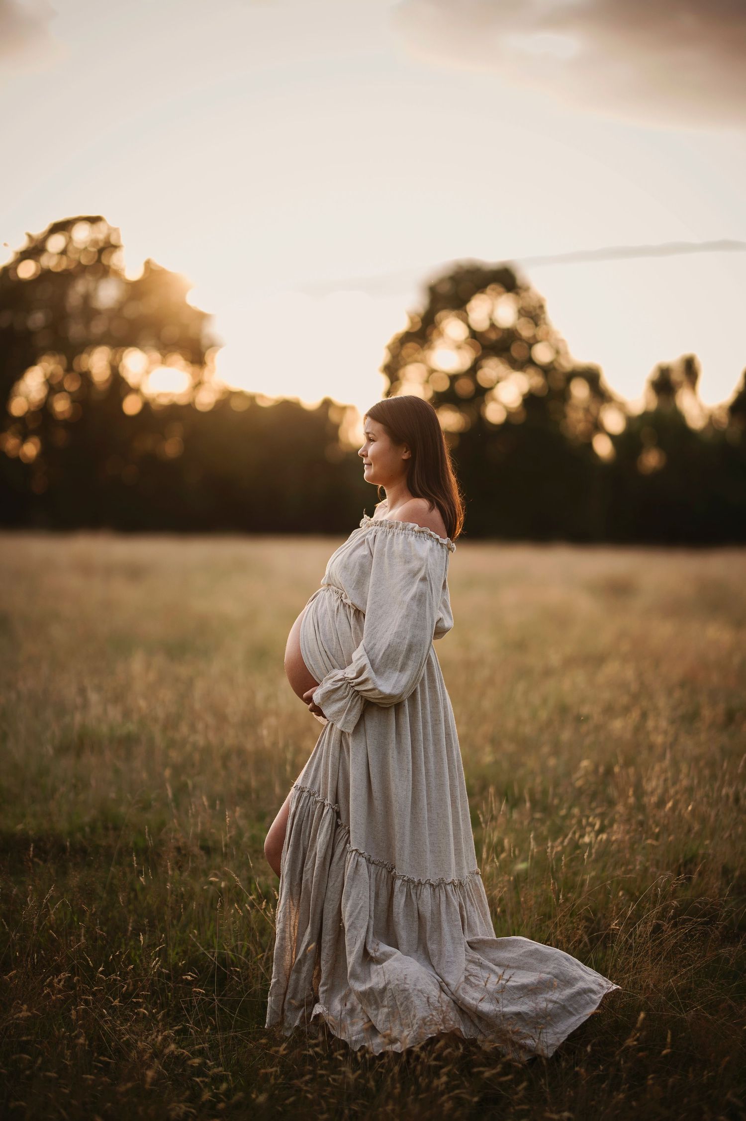 A maternity photo in a flowing white dress taken in a golden field at sunset.