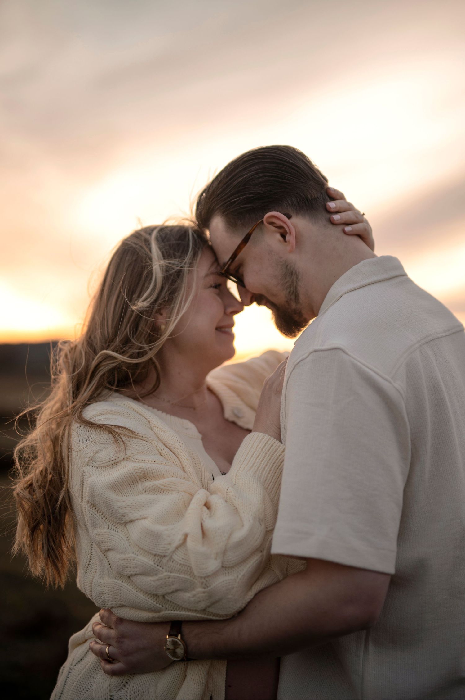 Romantic couple sharing an intimate moment at sunset, foreheads touching, wearing cream outfits with warm golden sky backdrop.