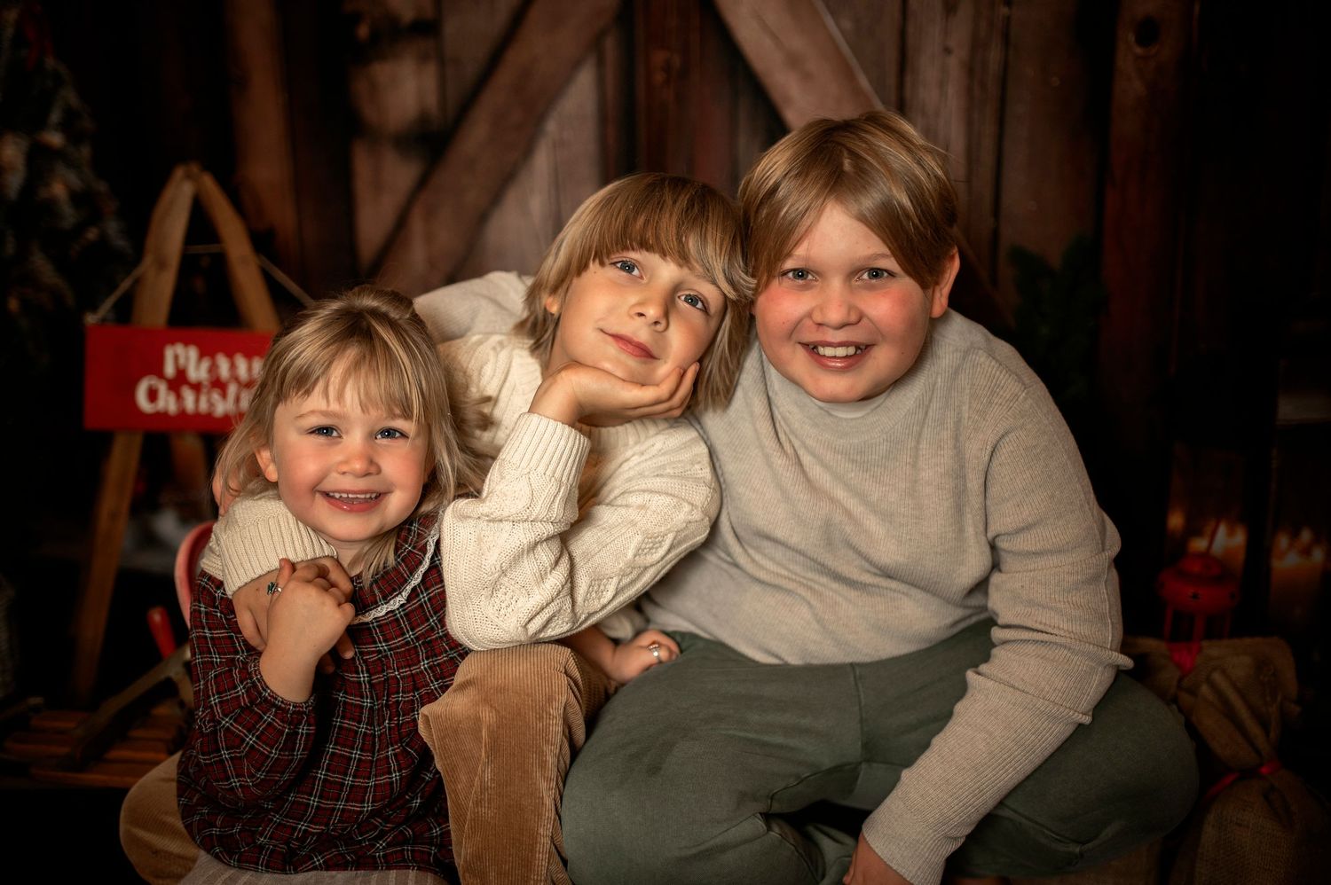 Three children pose together for a festive Christmas portrait in a rustic barn setting with holiday decorations.