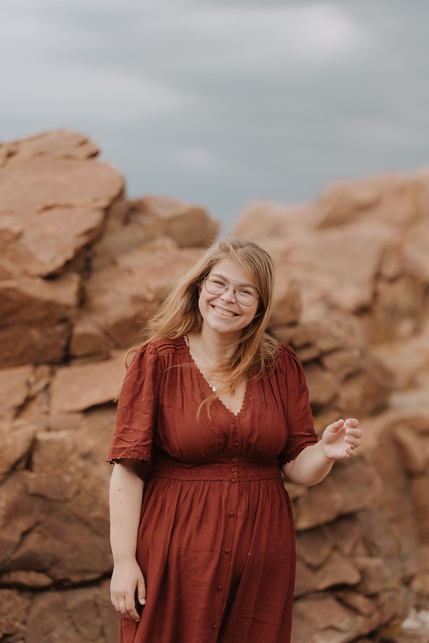 A smiling young woman with glasses in a red dress stands before rusty red rock formations under a cloudy sky.