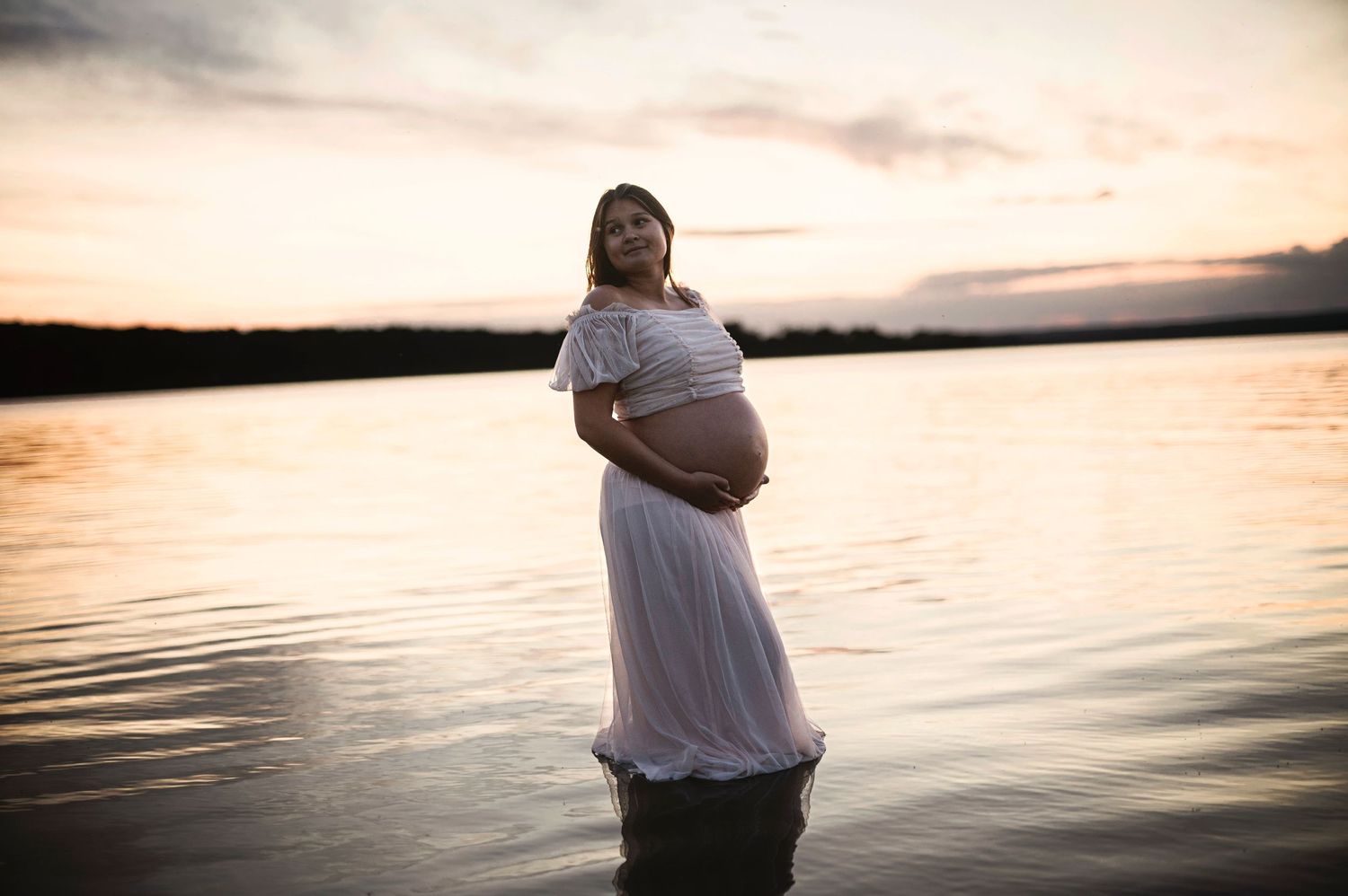 Silhouette of expectant mother in white dress standing in lake water during golden sunset.