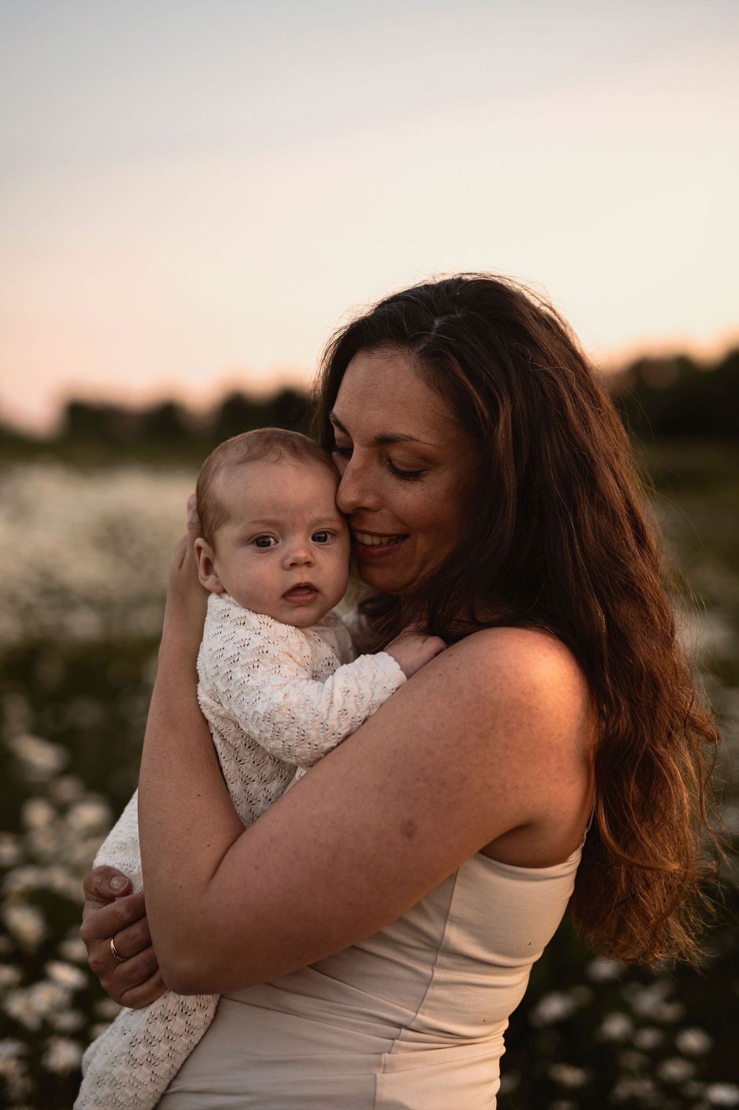 Nyföddfotografering i Skåne med mamma som håller sitt barn i solnedgång i blommande äng.