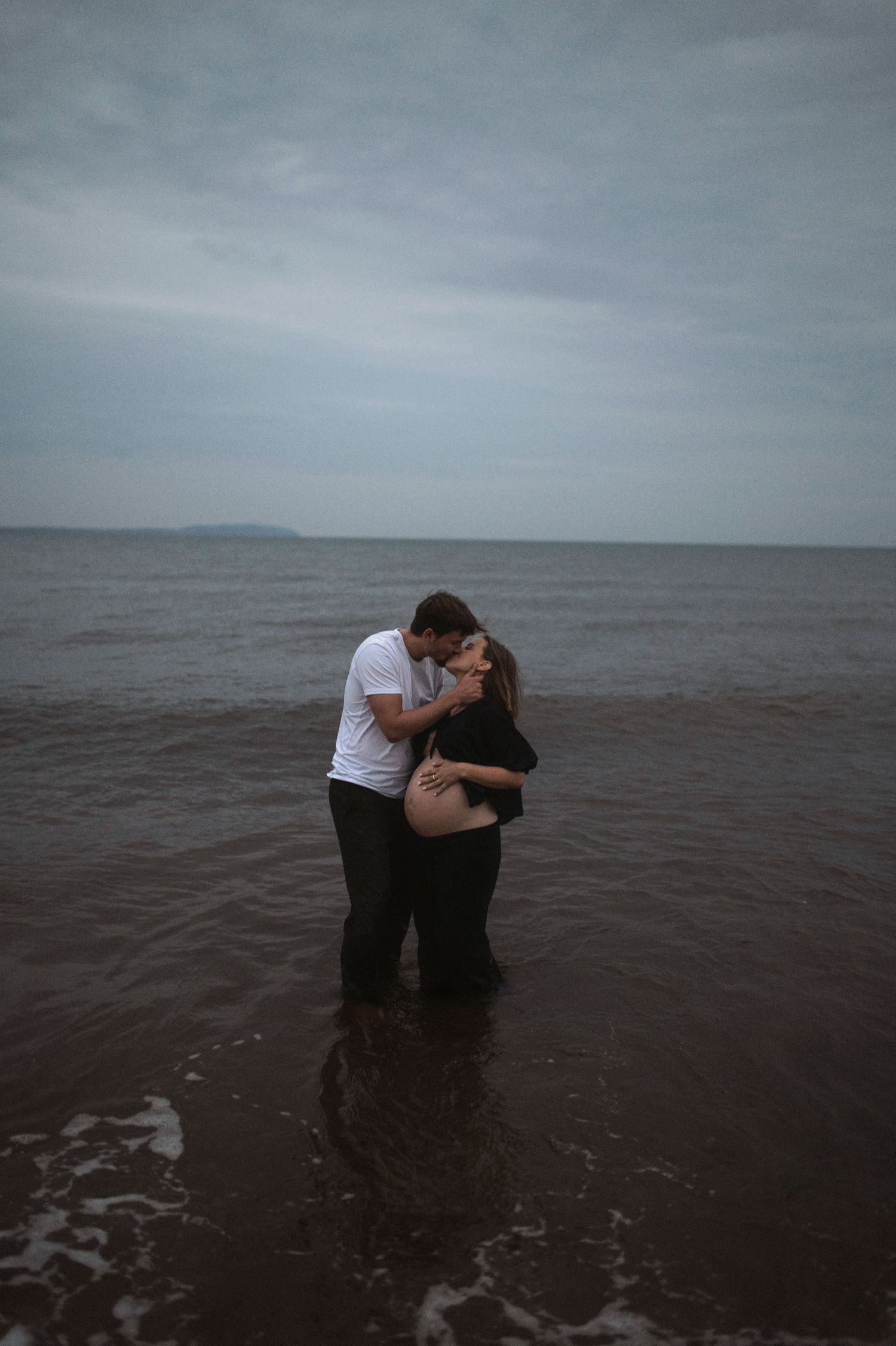 Cinematisk Gravidfotografering i Skåne med blivande föräldrar som kysser varandra i havet i dramatisk molnig himmel.