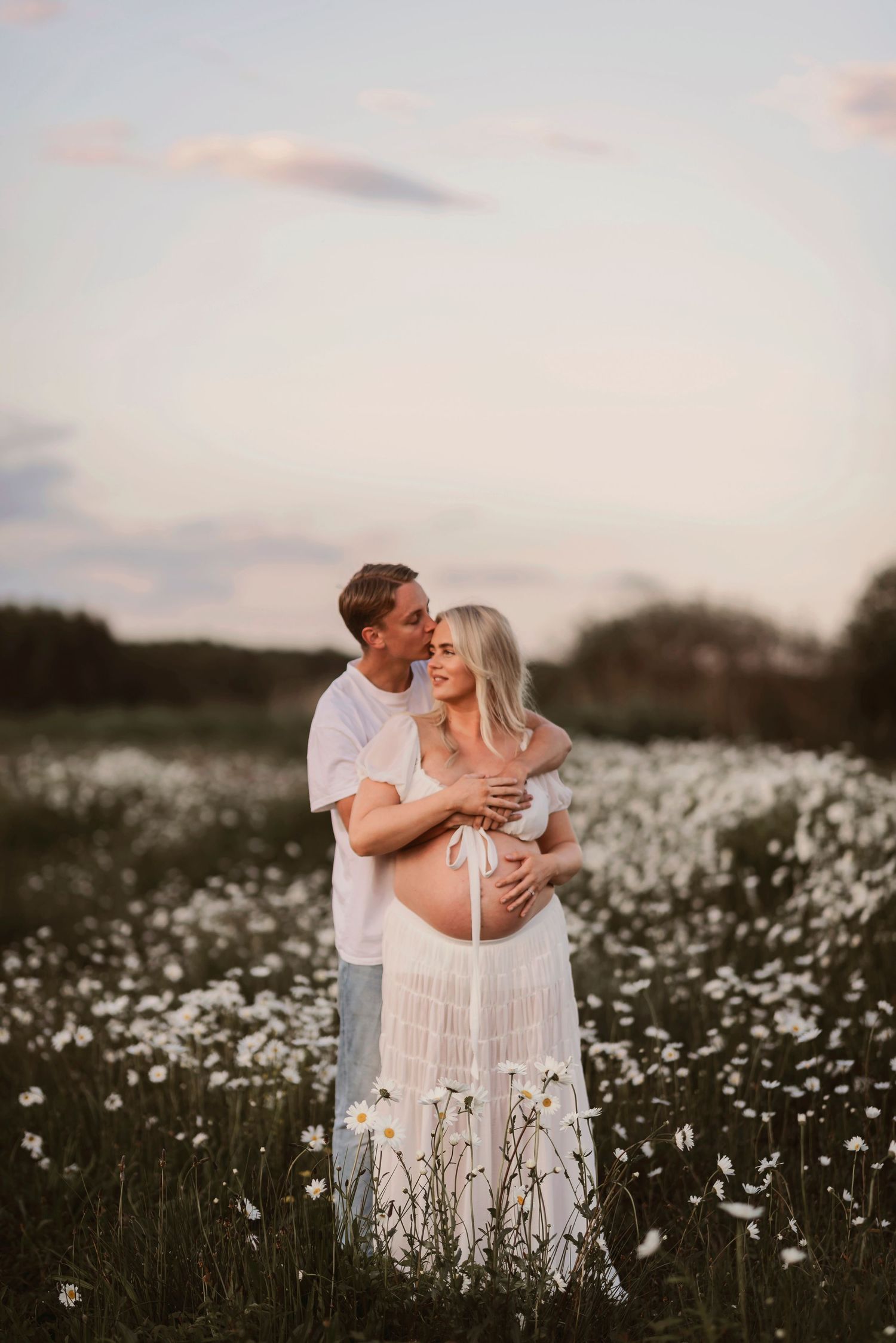 Gravidfotografering i Skåne med blivande föräldrar i blomsteräng vid skymning i mjukt kvällsljus.