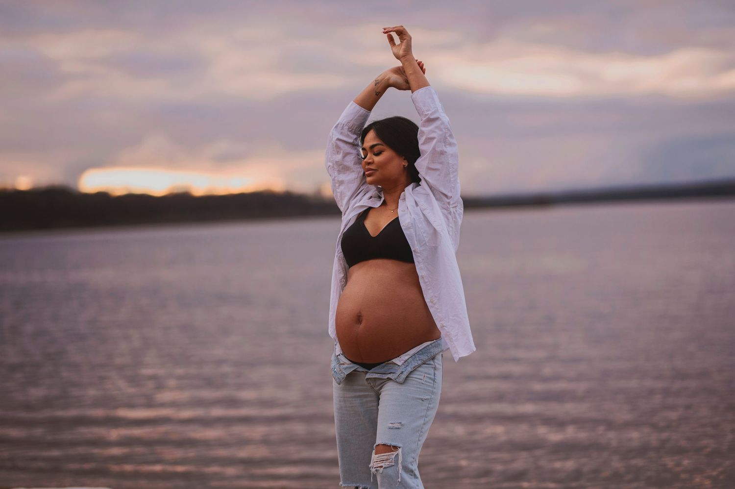 A pregnant figure in casual wear stretches by a lake at sunset with arms raised overhead.