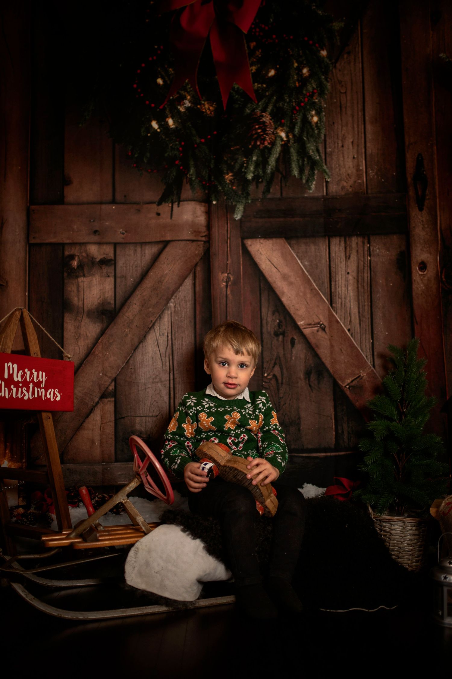 Young boy in Christmas sweater sits on sled before rustic barn door with holiday wreath and Merry Christmas sign.