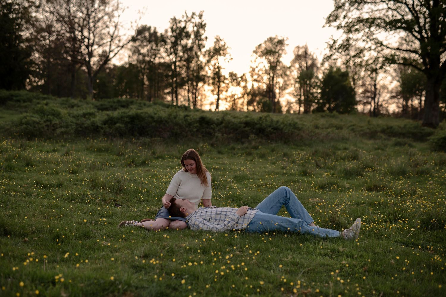 Couple relaxing in a green meadow at sunset, woman sitting while man lies down among wildflowers.