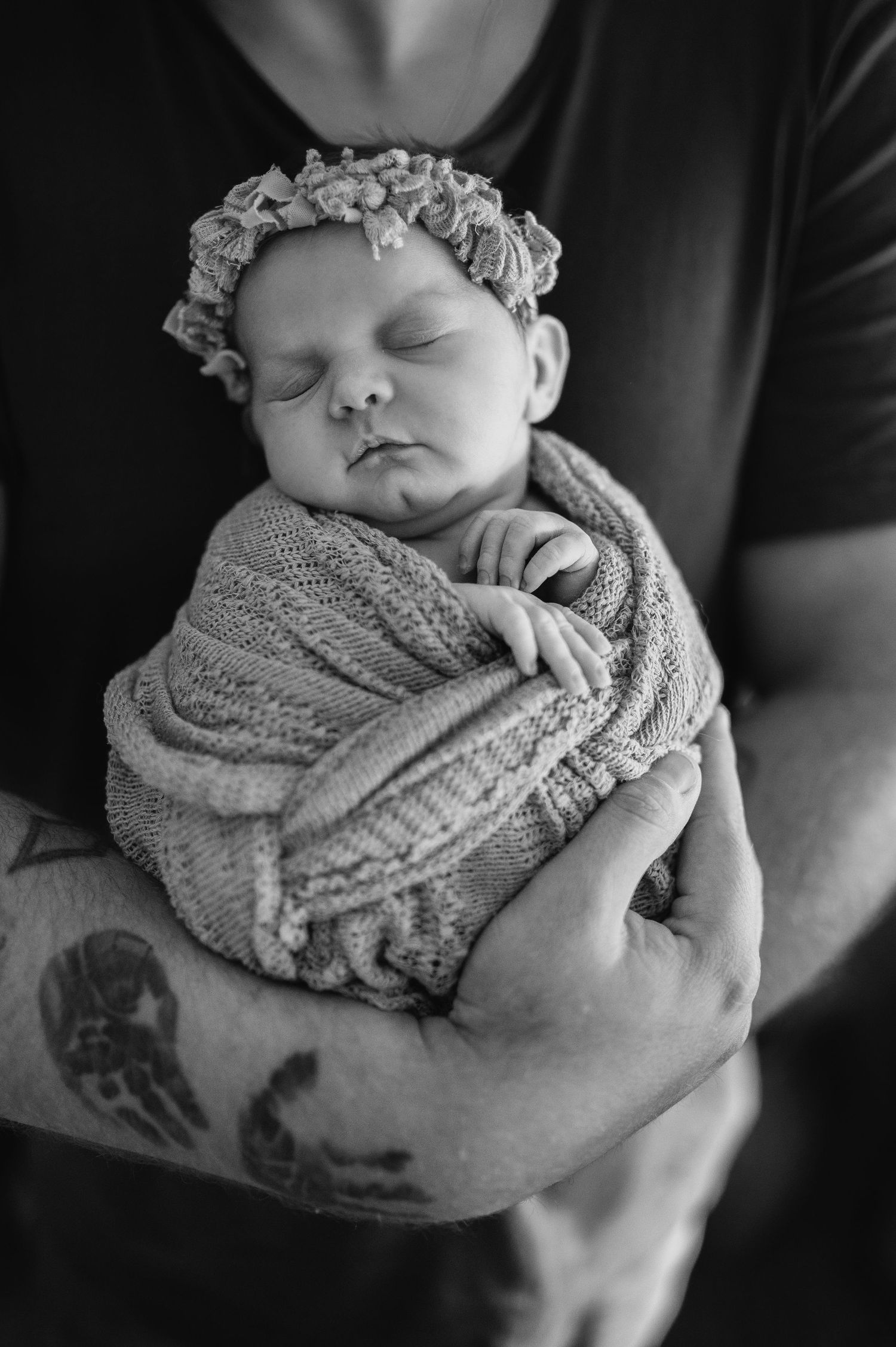 A sleeping newborn wrapped in gray knit fabric with a floral headband rests peacefully in gentle arms.