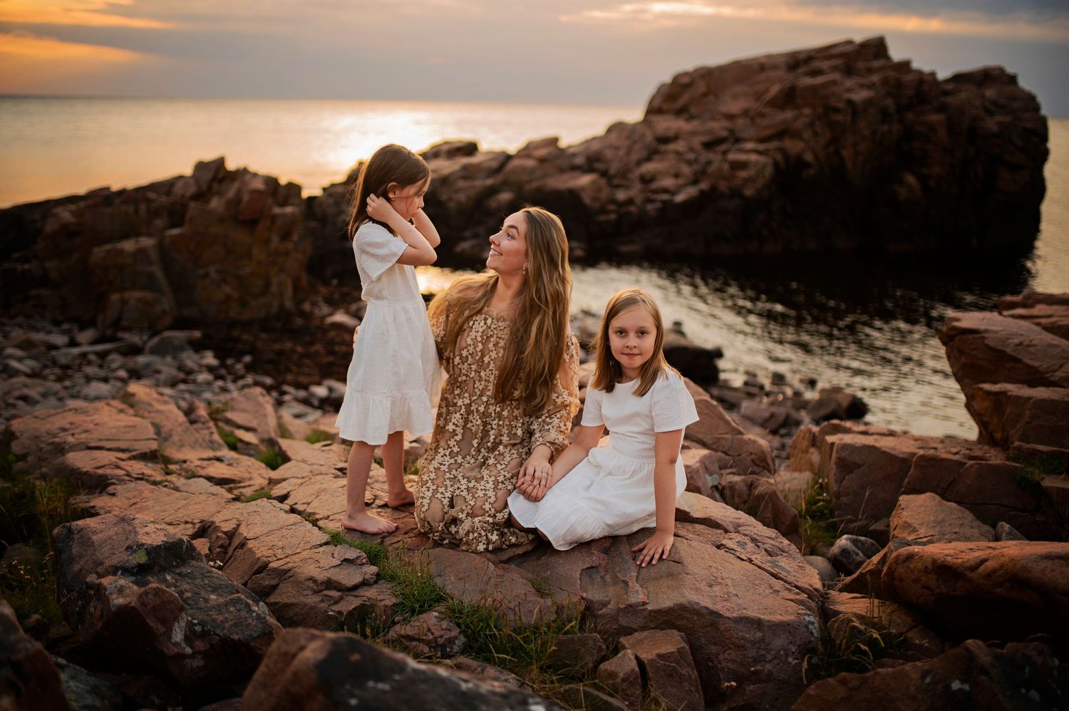 Sunset photoshoot on rocky coastal shore with white dresses and golden light.