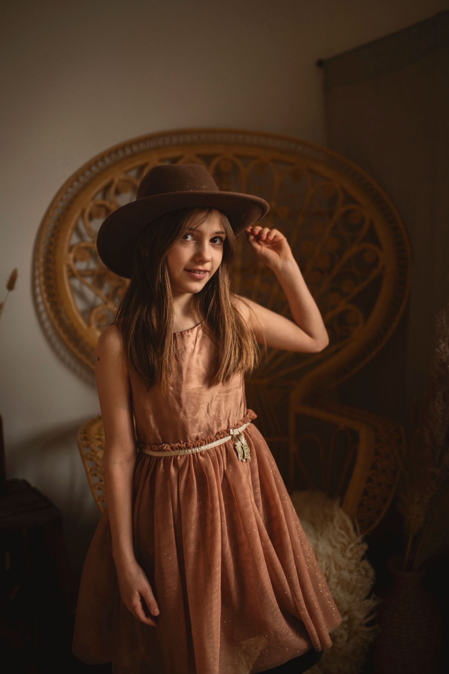 Boho-styled portrait features a brown dress and wide-brim hat against a peacock chair.