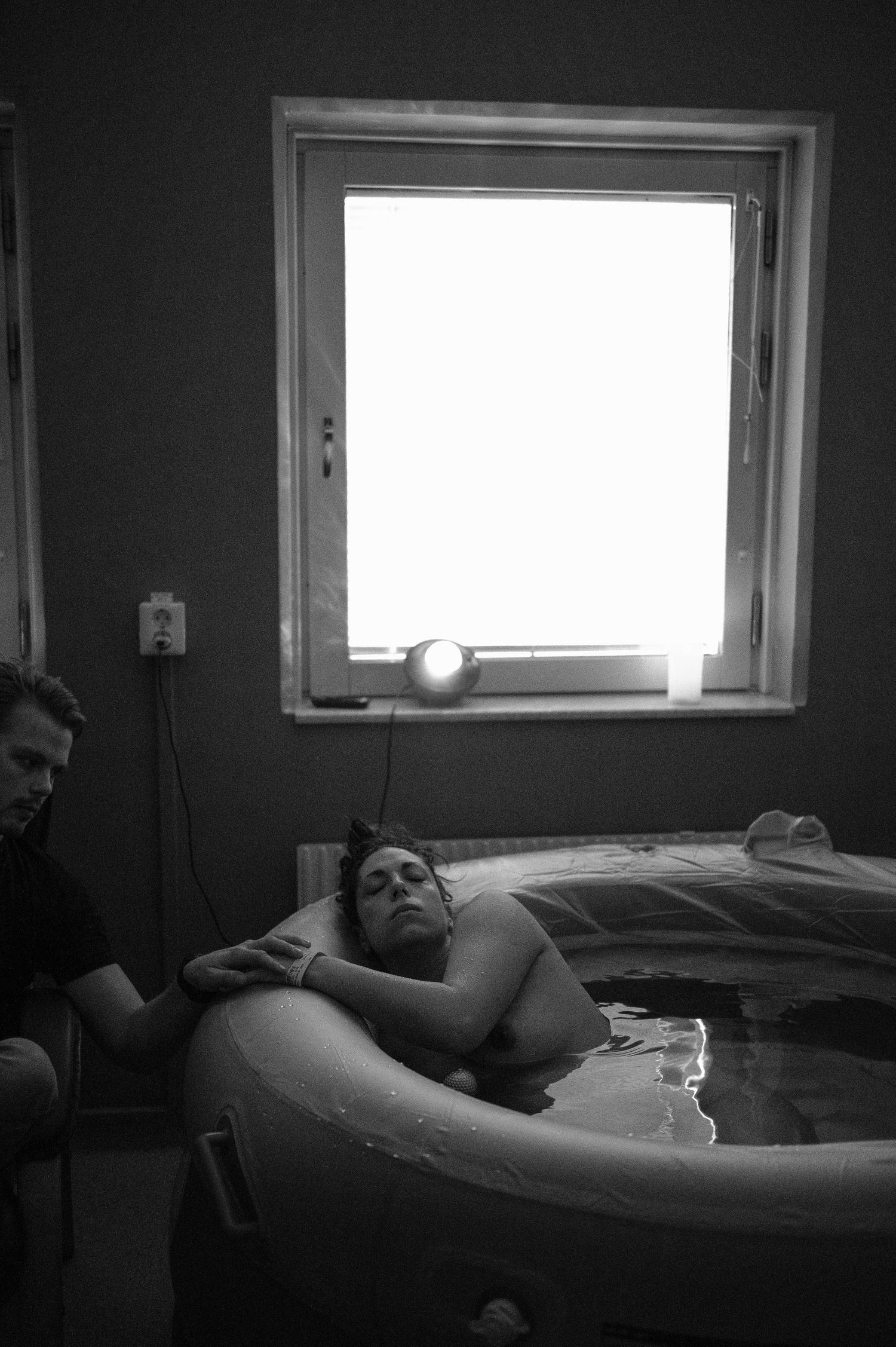 A person labors in a birthing pool near a bright window during childbirth while supported by their partner.