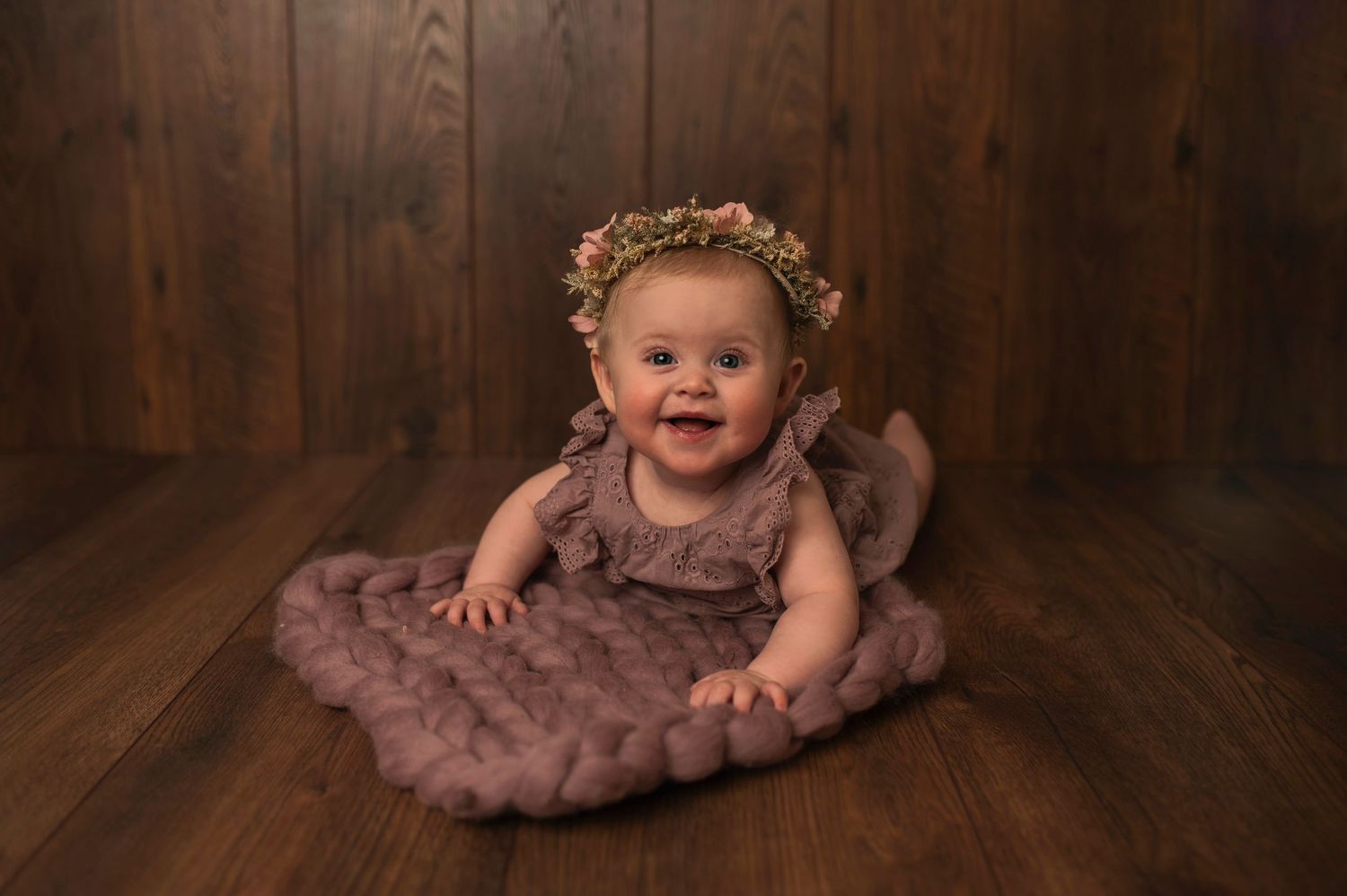 Baby in mauve dress and floral crown sitting on wooden floor.