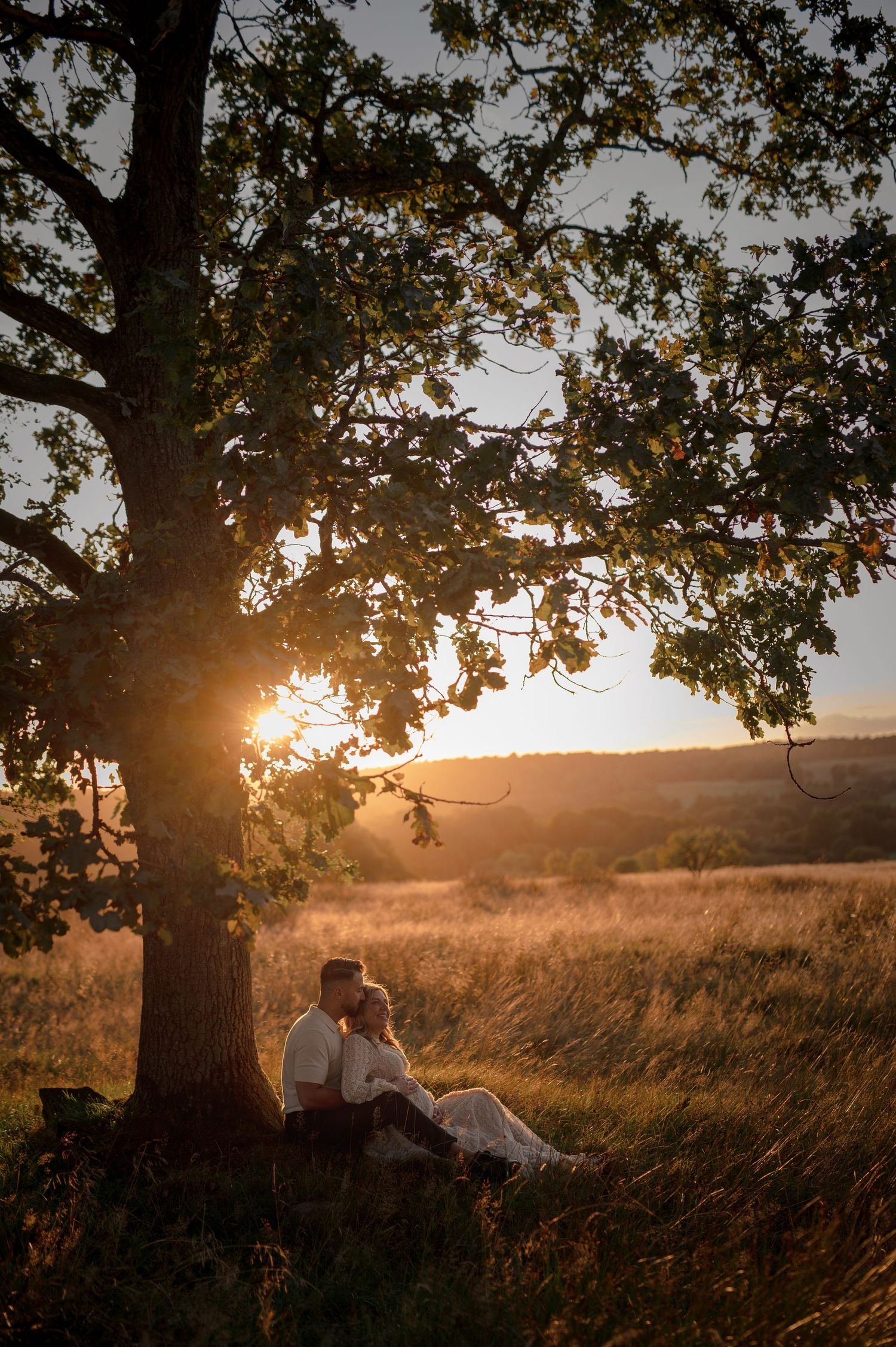 Couple sitting under a large tree at golden sunset in a scenic meadow landscape.