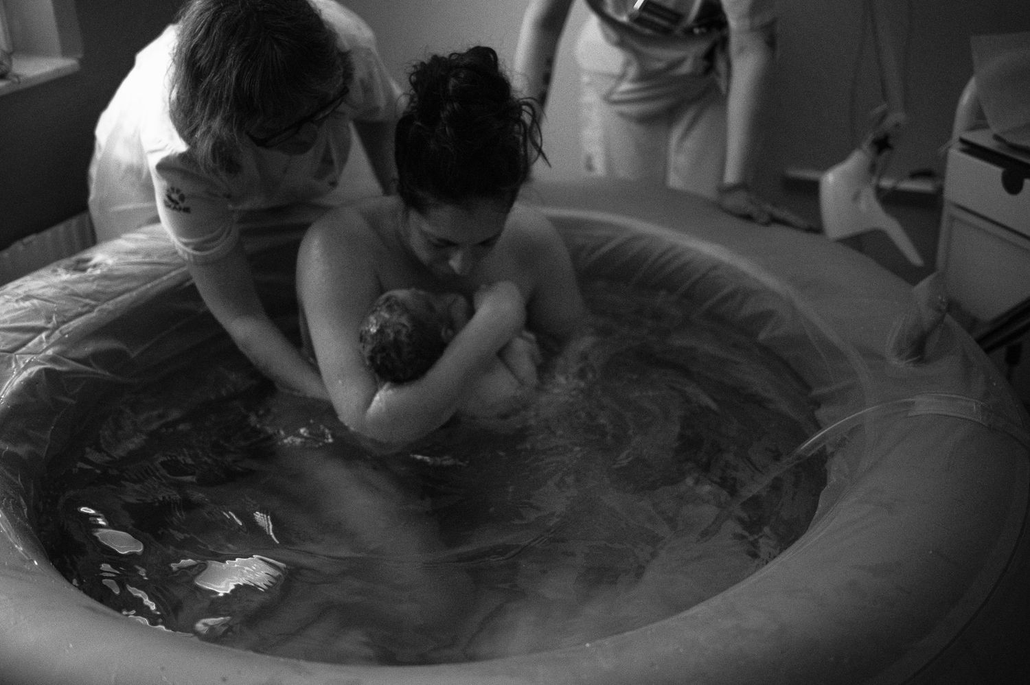 A black and white photograph shows a water birth in progress with people attending in a birthing pool.