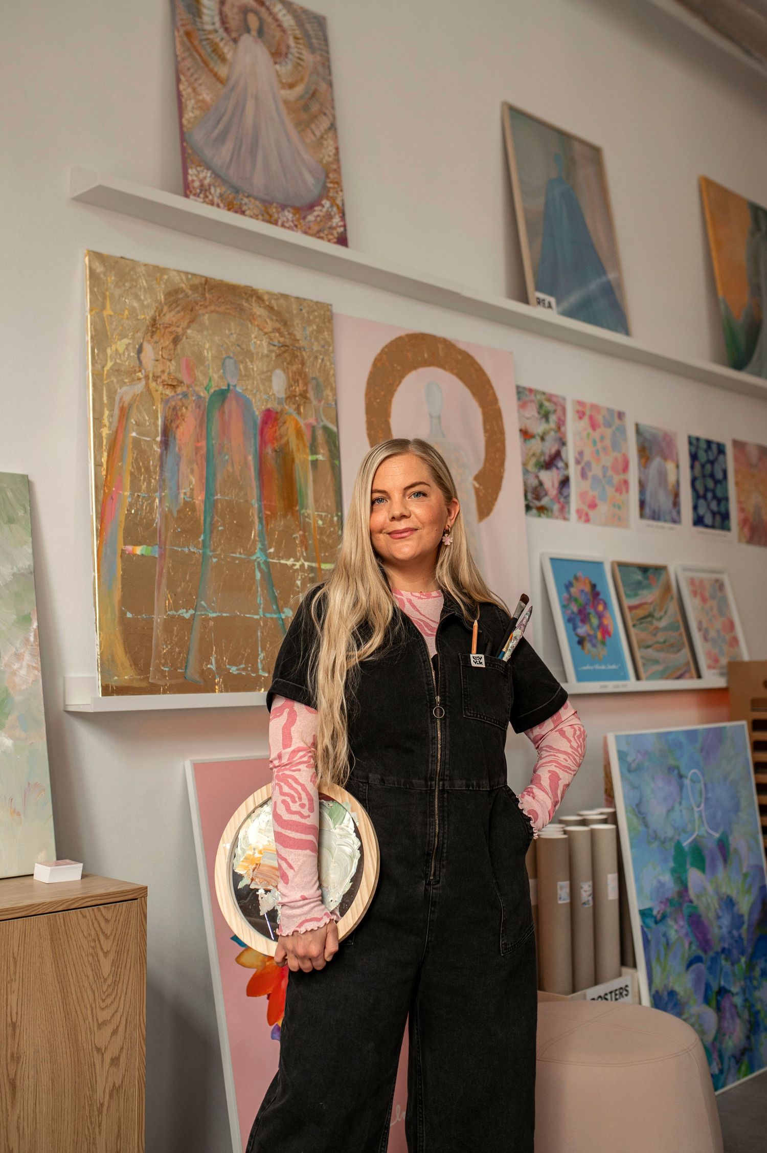 Female artist in black overalls holding embroidery hoop standing in colorful art studio with paintings.