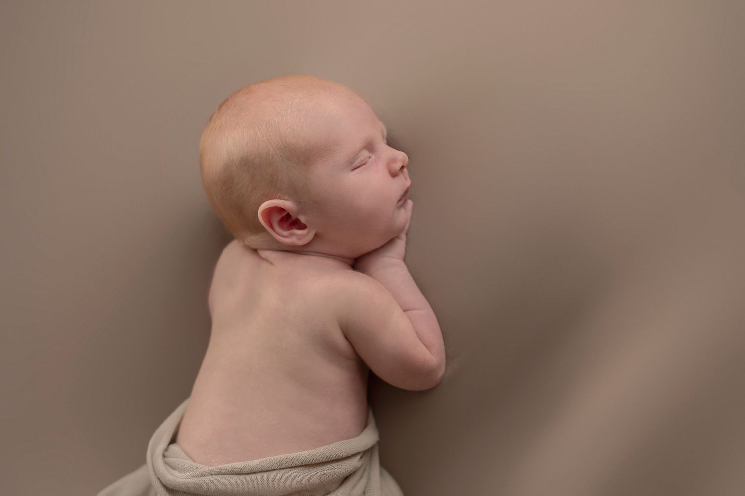 Sleeping newborn posed on side against neutral brown background.