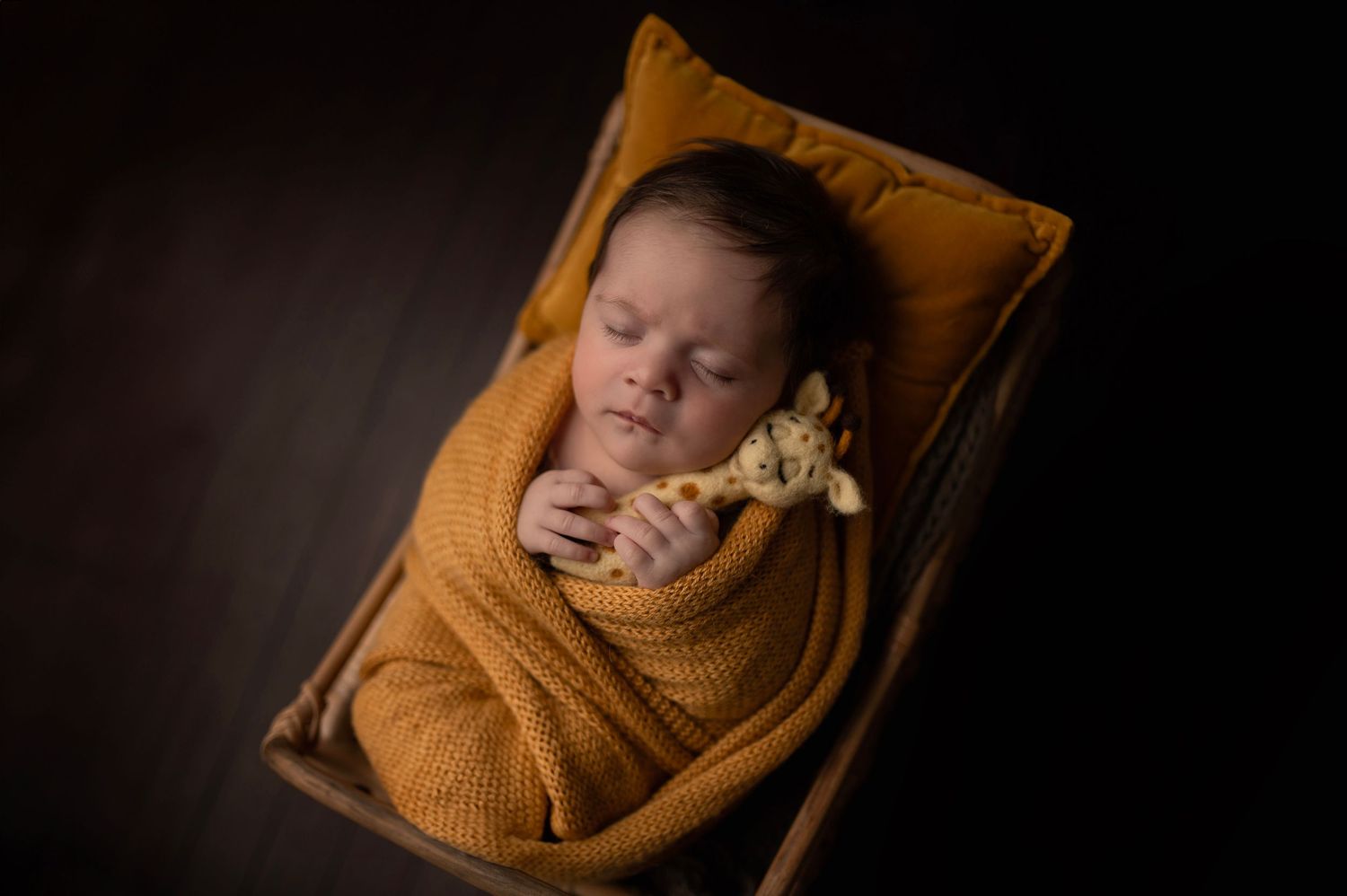 Peaceful newborn baby swaddled in mustard colored wrap on wooden bed.