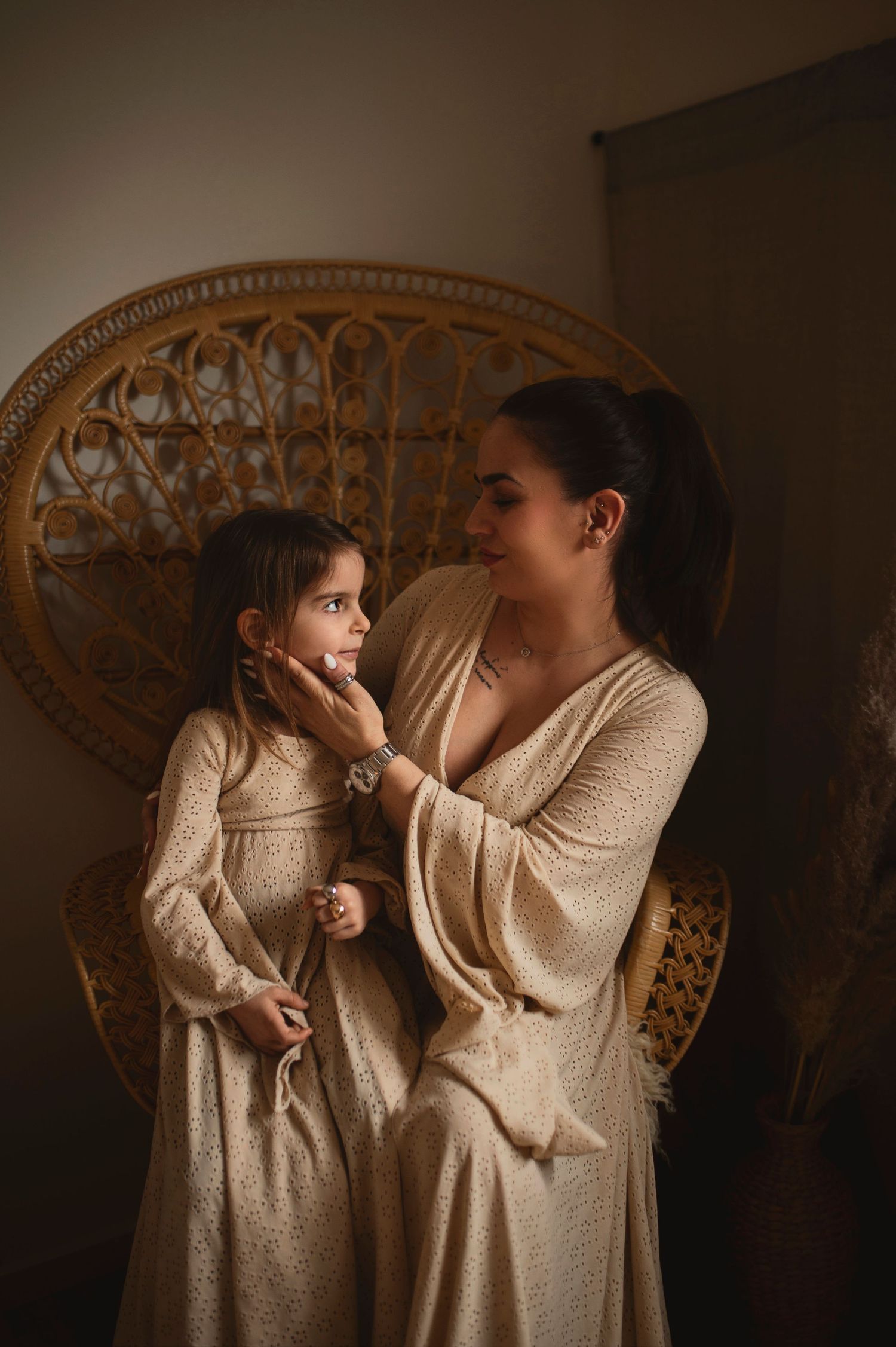 Two people in cream-colored dresses share a tender moment against a wicker chair backdrop.