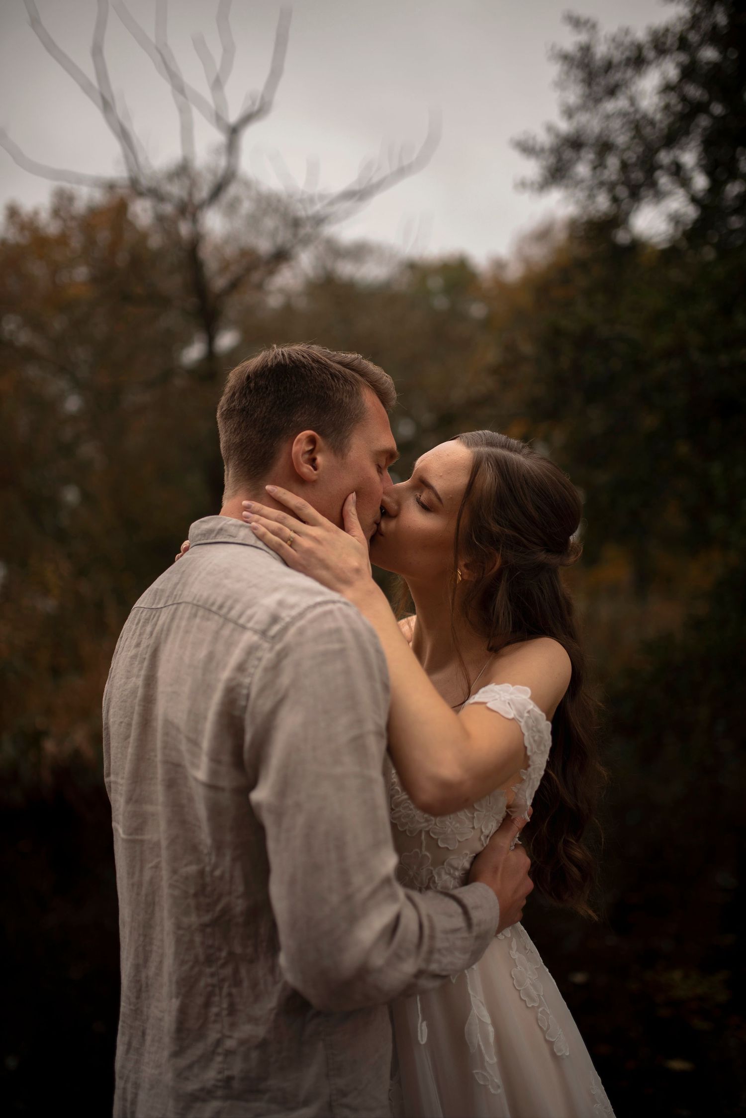 Couple sharing a romantic kiss outdoors in autumn, bride in white lace gown embracing groom in gray linen shirt.