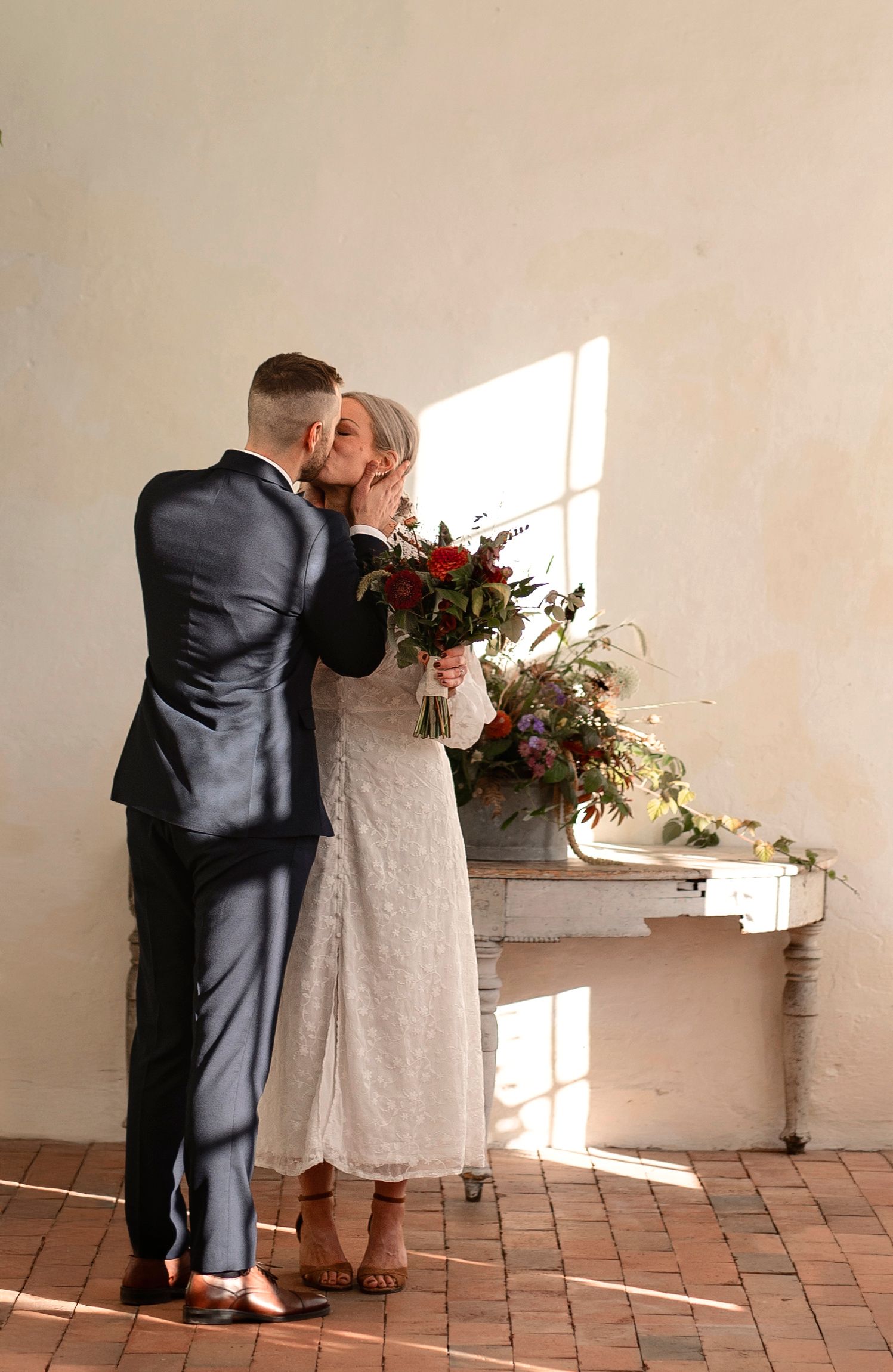 Bride and groom sharing a kiss near a sunlit window, bride holding a lush red and green floral bouquet in a rustic venue.