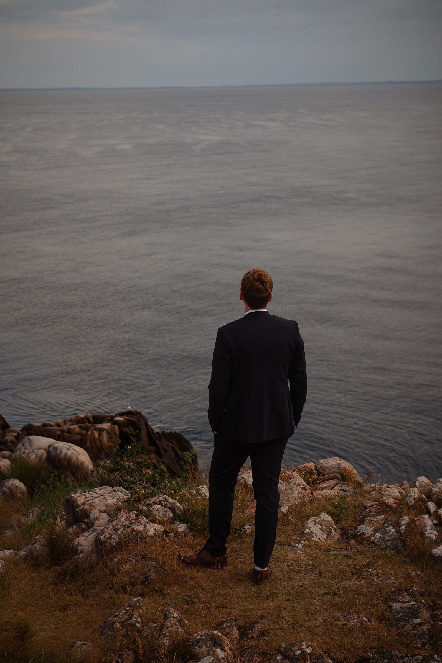 Man in dark suit standing on rocky cliff edge overlooking calm gray ocean waters. at a Kullaberg Elopment