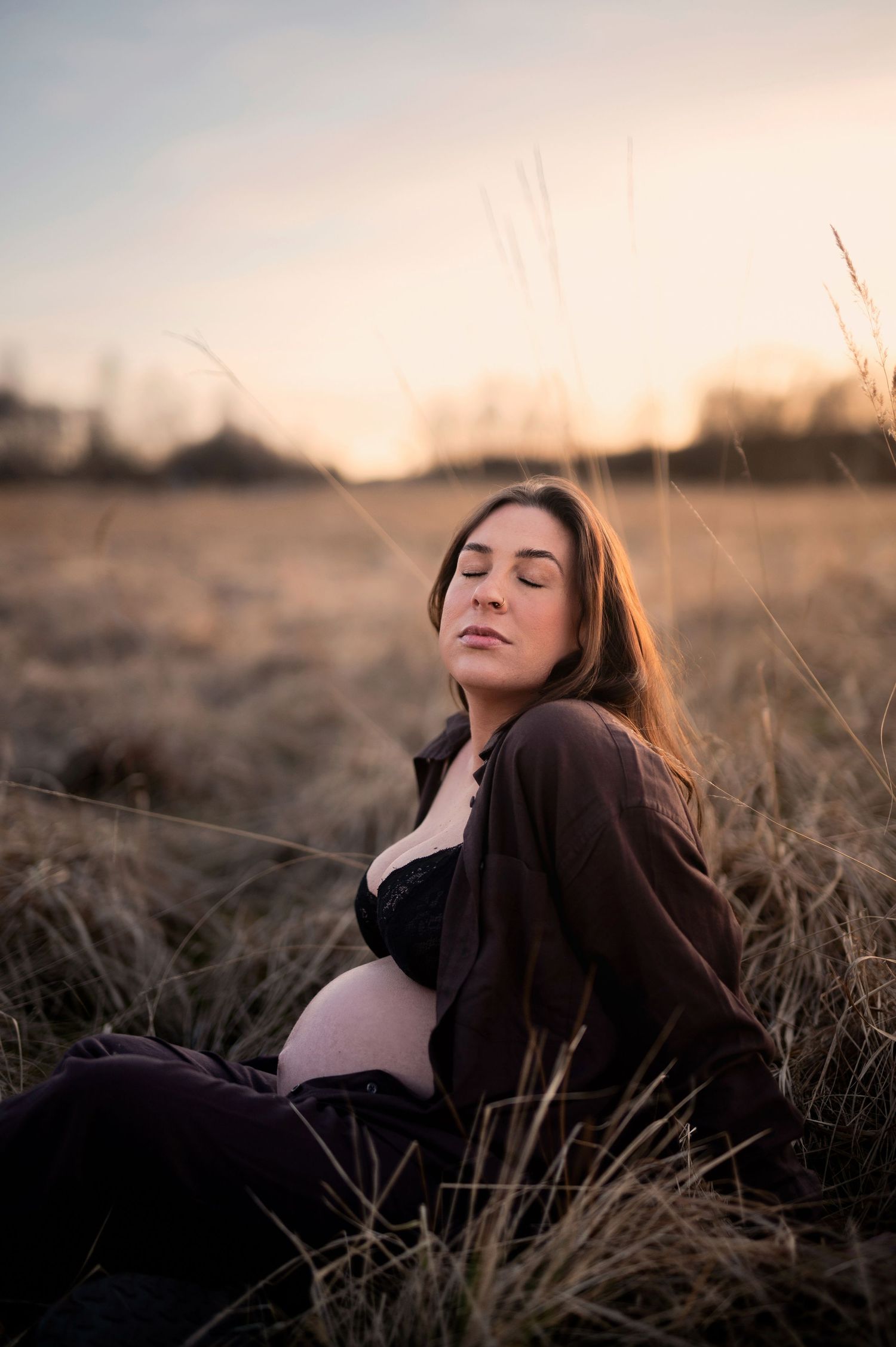 Gravid kvinna sitter i ett gyllene fält i solnedgången med slutna ögon och mörka kläder, gravidfotografering i Skåne
