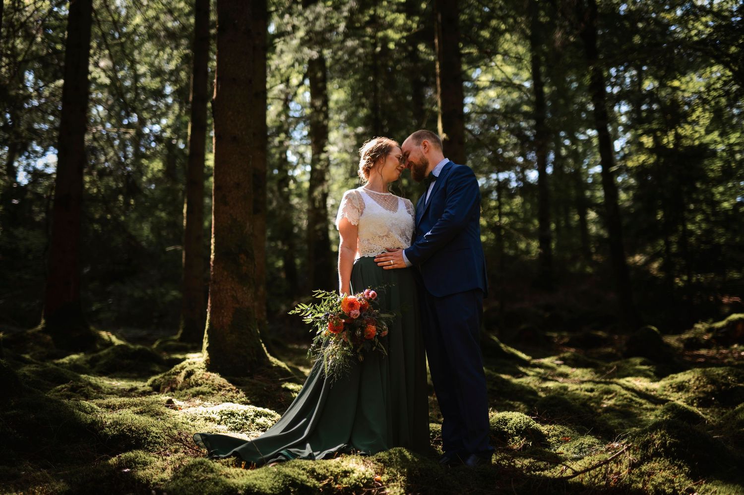 Bride and groom share romantic moment in sunlit mossy forest, bride wearing green gown holding red bouquet. in Falkenberg in Halland