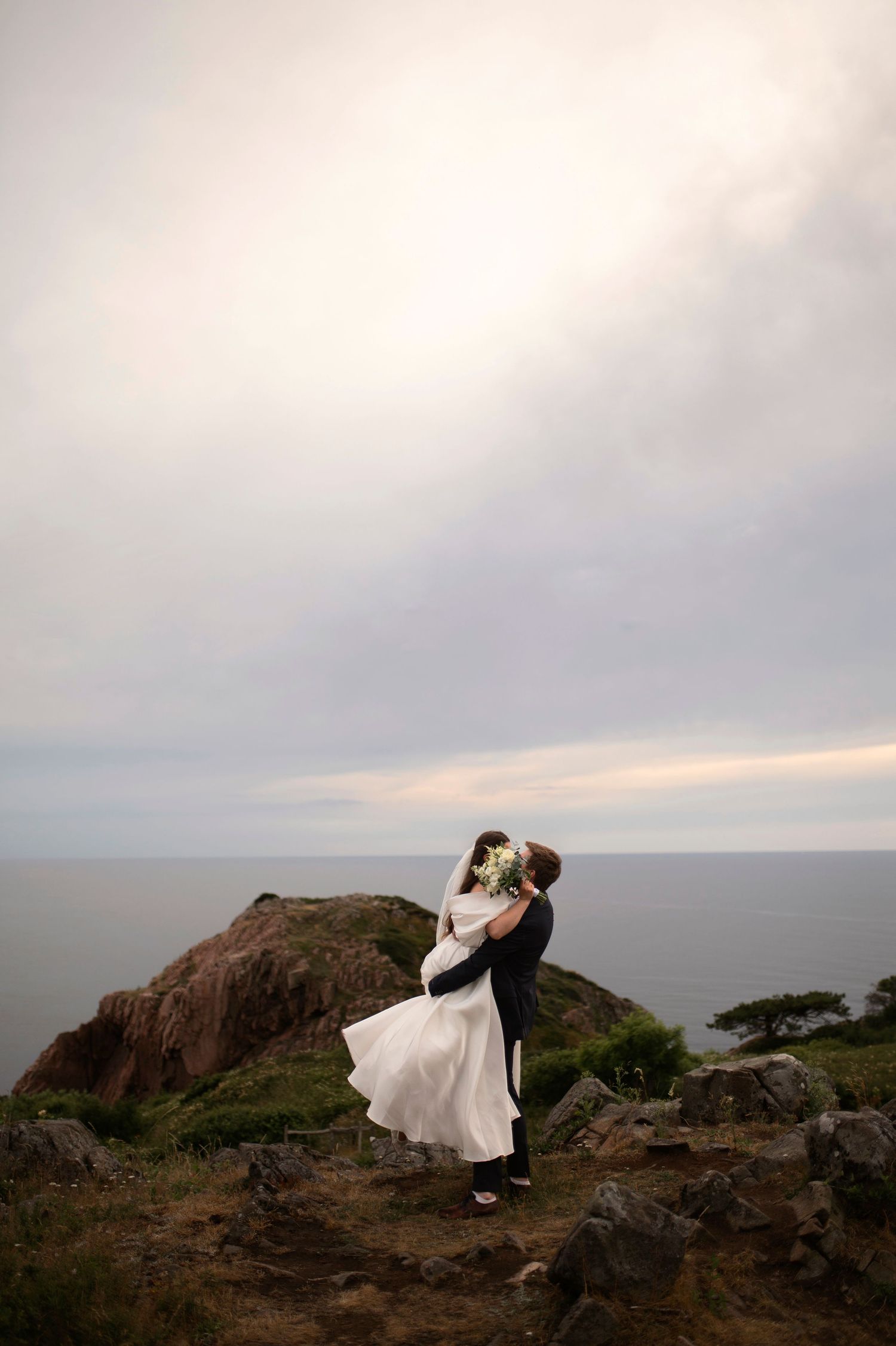 Groom lifts bride in white dress on rocky coastal cliff with dramatic cloudy sky and ocean backdrop at a Elopement in Mölle in Skåne